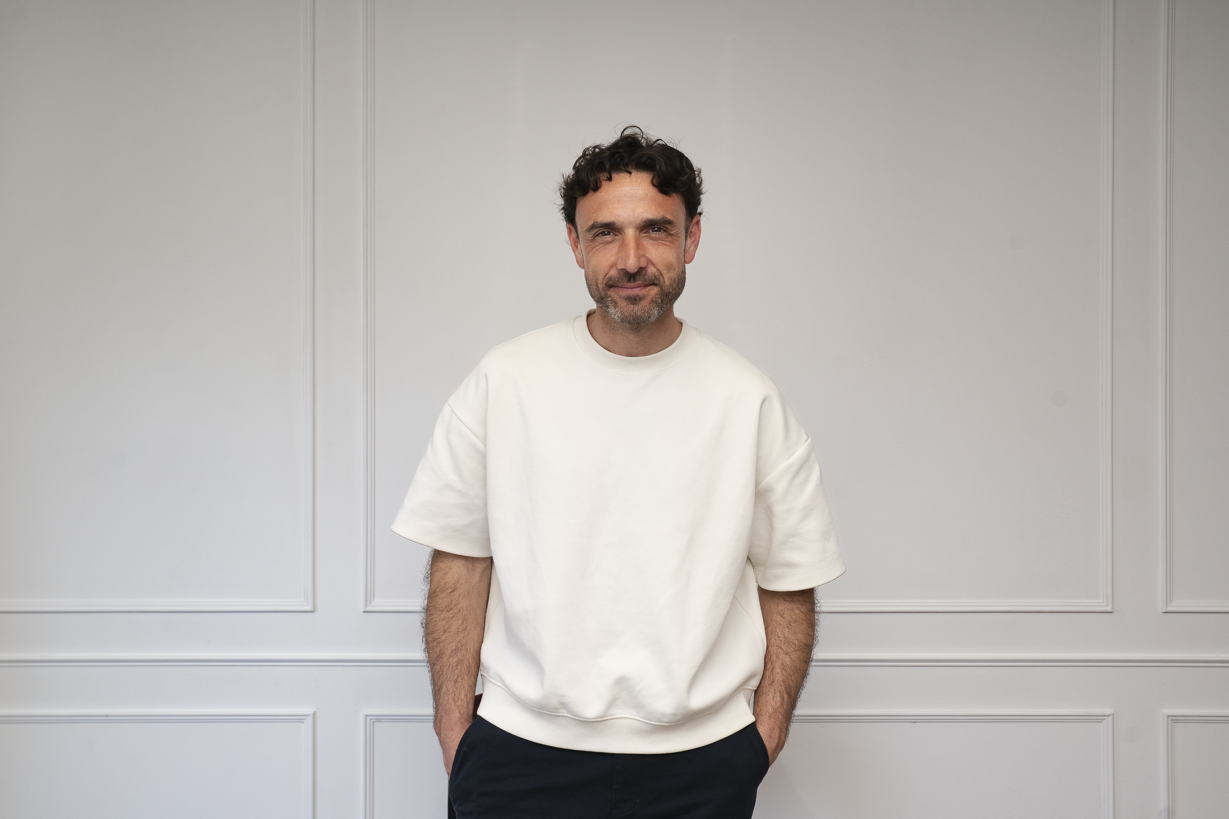 A man with dark curly hair and a beard, wearing a white T-shirt and dark pants, standing against a white paneled wall, looking at the camera with a slight smile.