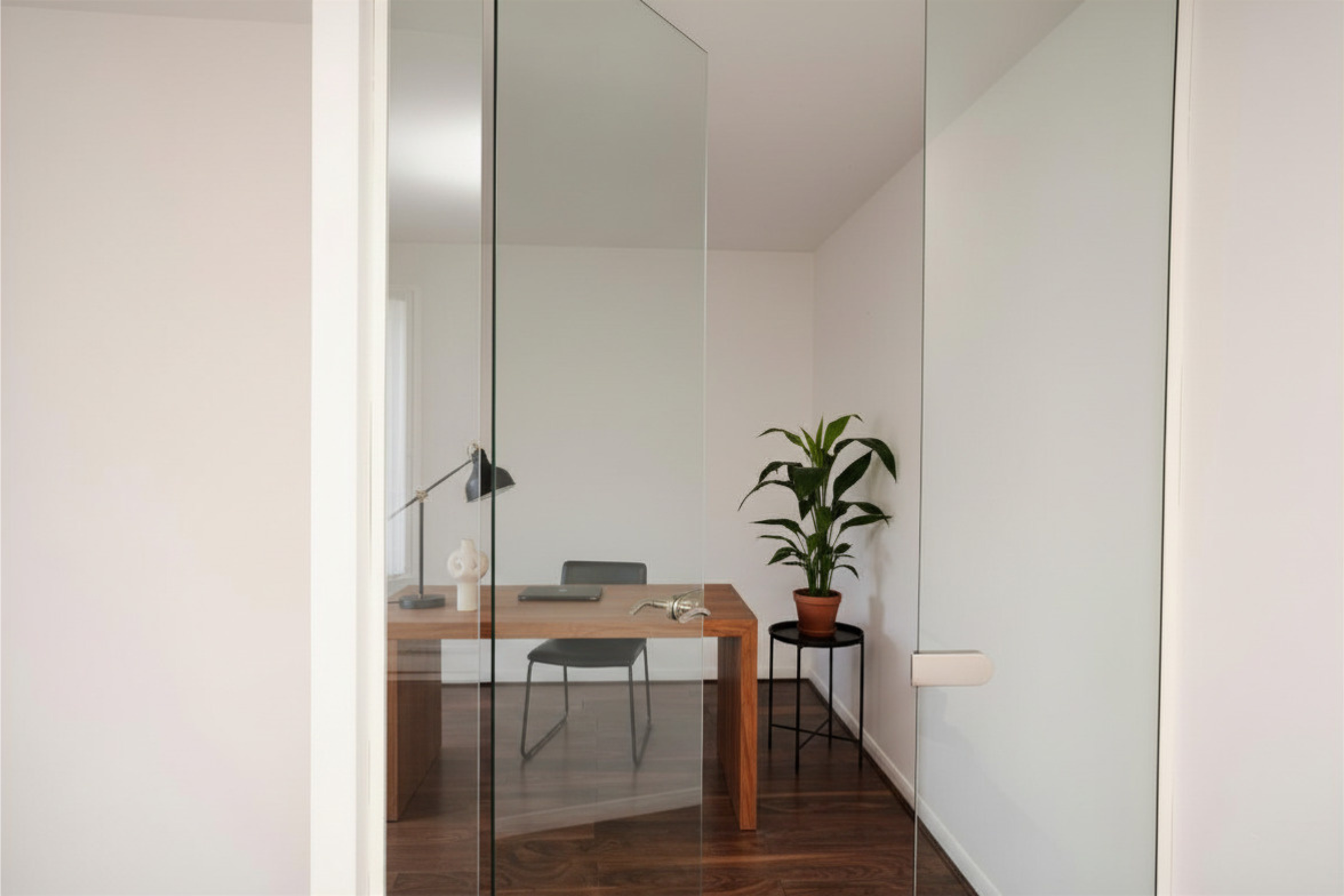 A mirror reflects a room with a wooden table, a chair, a potted plant, a desk lamp, a tablet, and a decorative figurine.