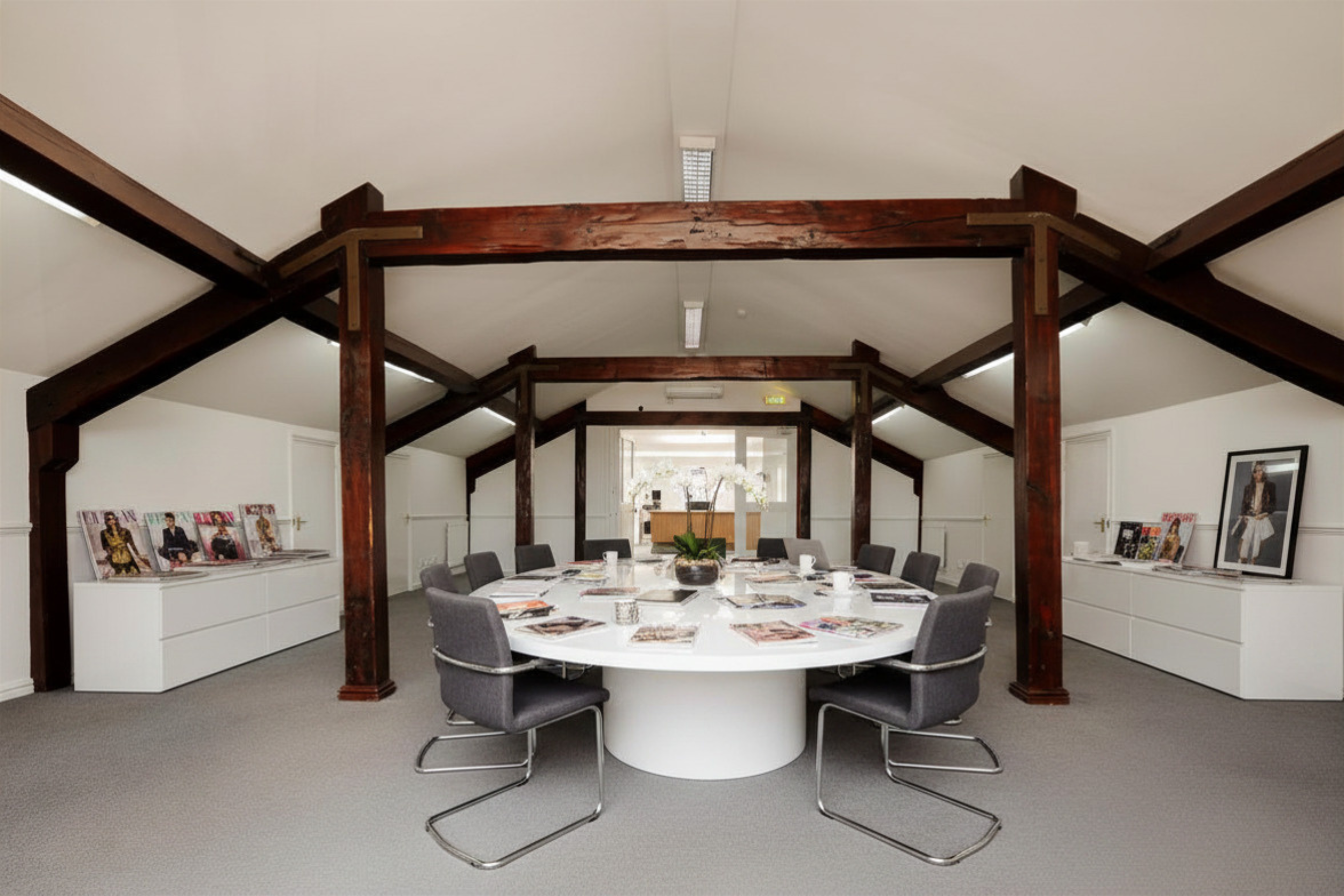 Conference room with a round white table, gray chairs, decorative magazines and framed pictures on white cabinets, rustic wooden beams on the ceiling.