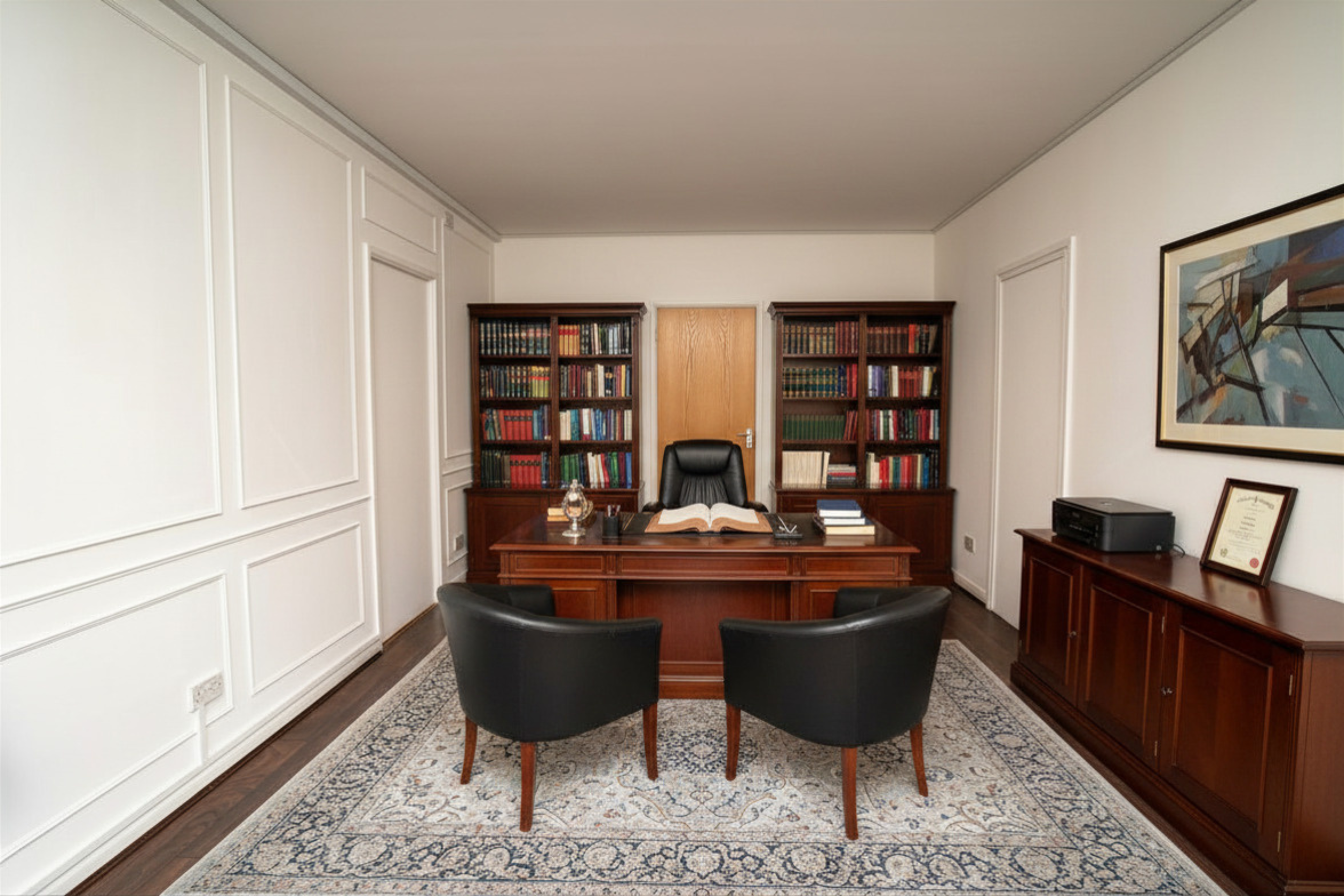 A professional office with a wooden desk, black leather chairs, and bookshelves filled with books. There's a painting on the wall, a framed certificate, a printer on a side table, and an open book on the desk.