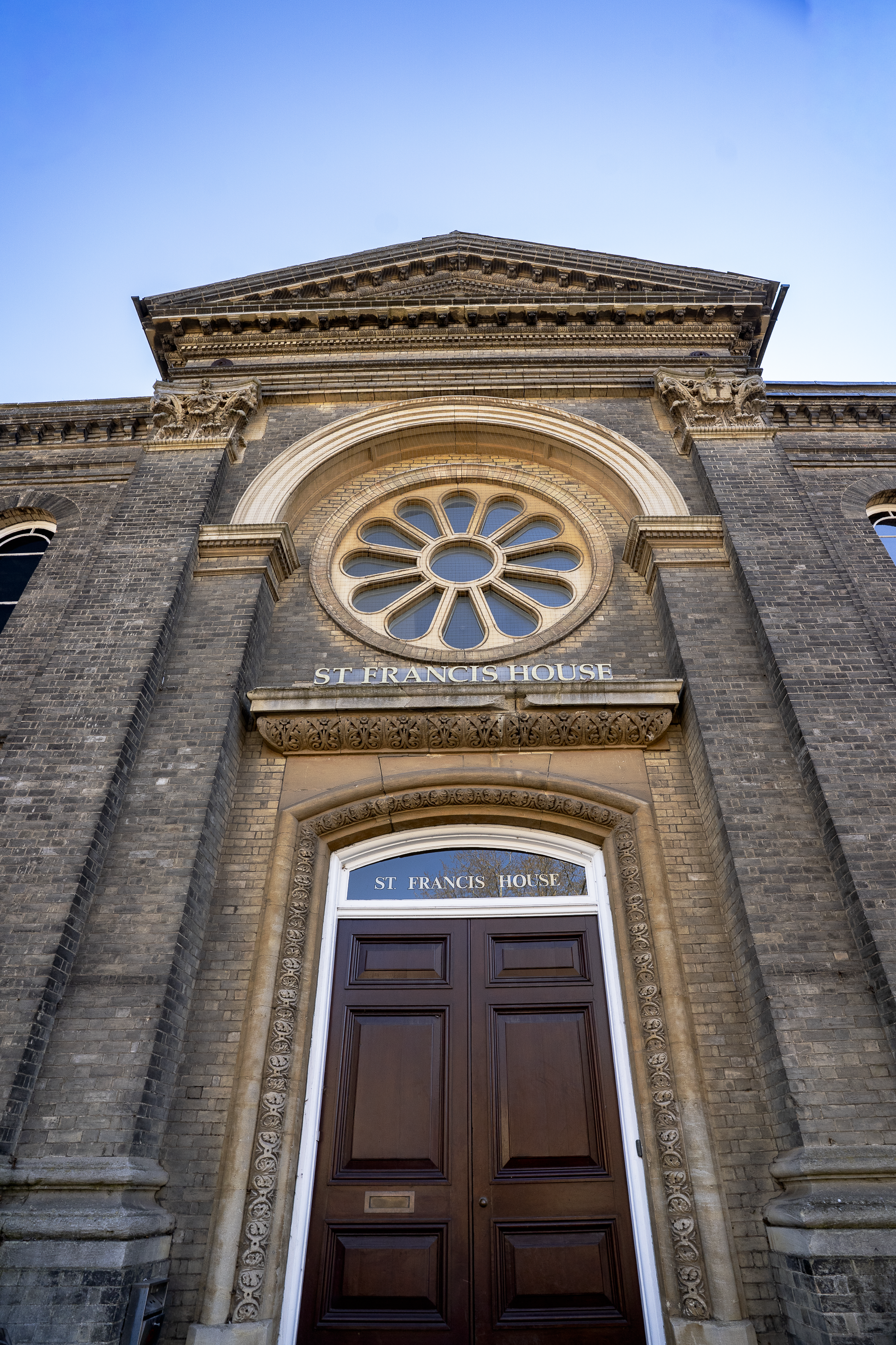 The front entrance of St. Francis House, a historic brick building with an ornate stone archway above the door, featuring a large circular window with decorative spokes.