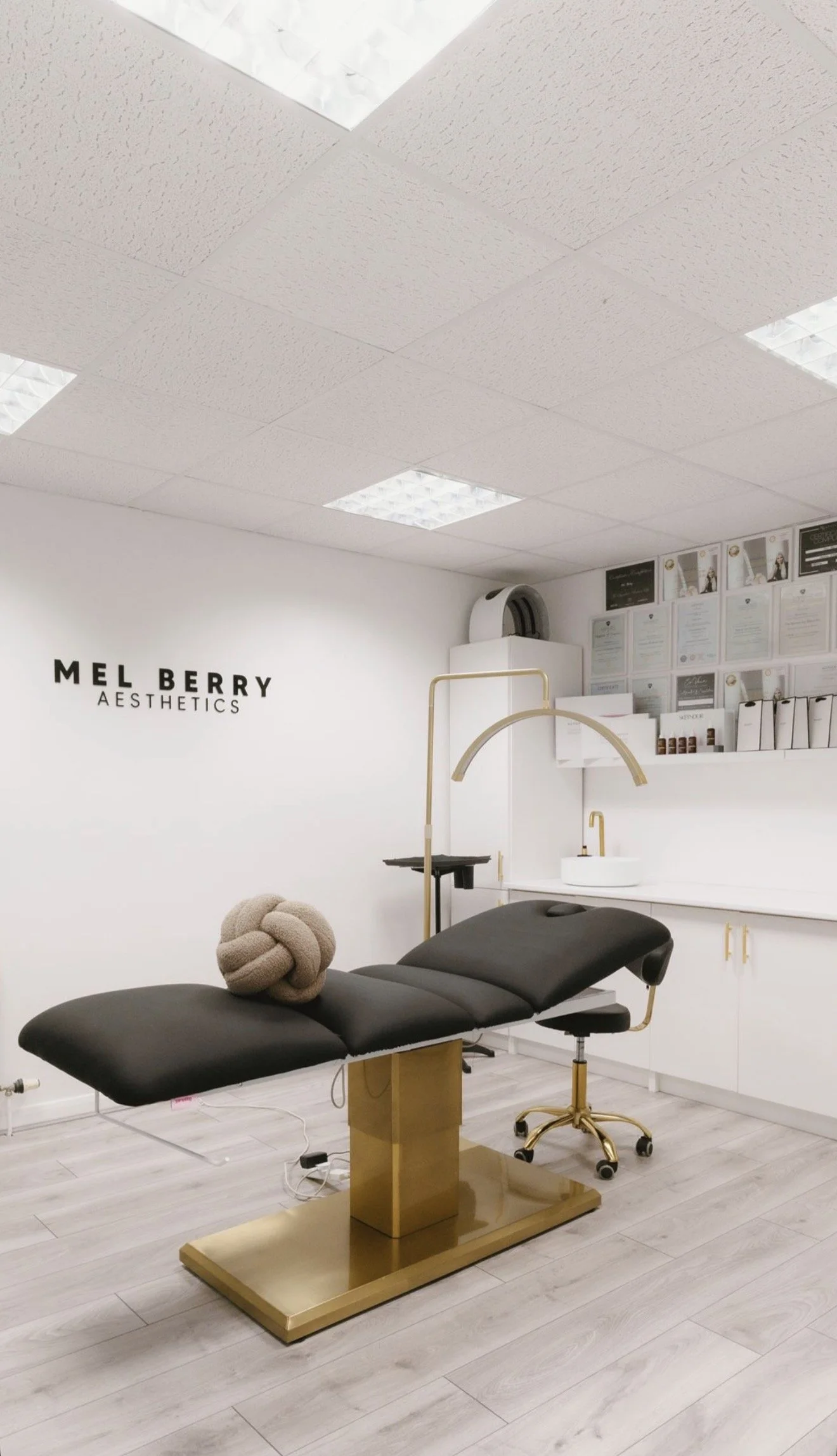 Interior of a skincare clinic or treatment room with a black massage or treatment table, a beige knot-shaped pillow, gold accents, and a white wall with a sign that reads "Mel Berry Aesthetics."