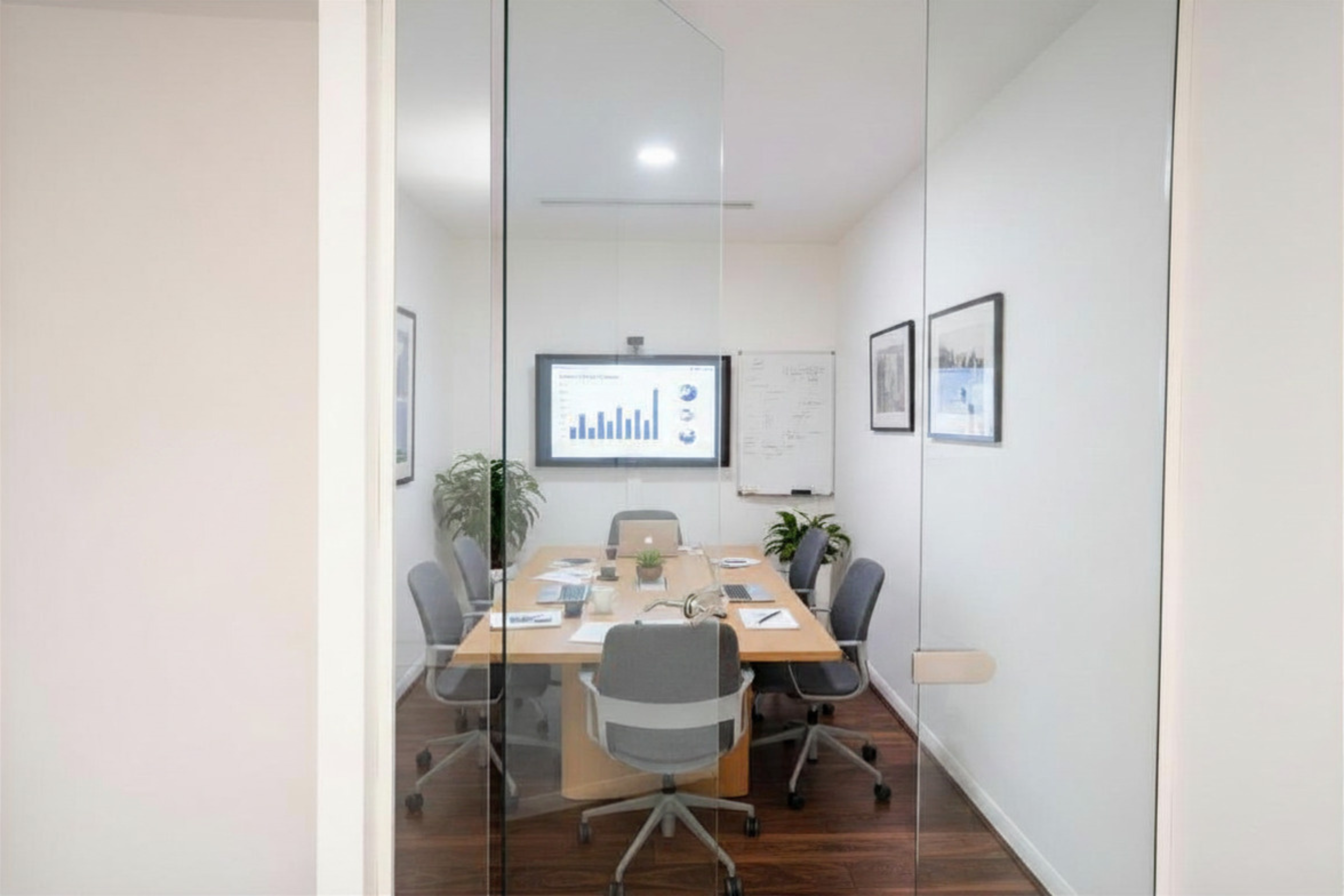 A conference room seen through glass walls, with a large wooden table surrounded by nine office chairs, some with laptops and papers on them. There is a wall-mounted screen displaying charts, and framed pictures on the wall.