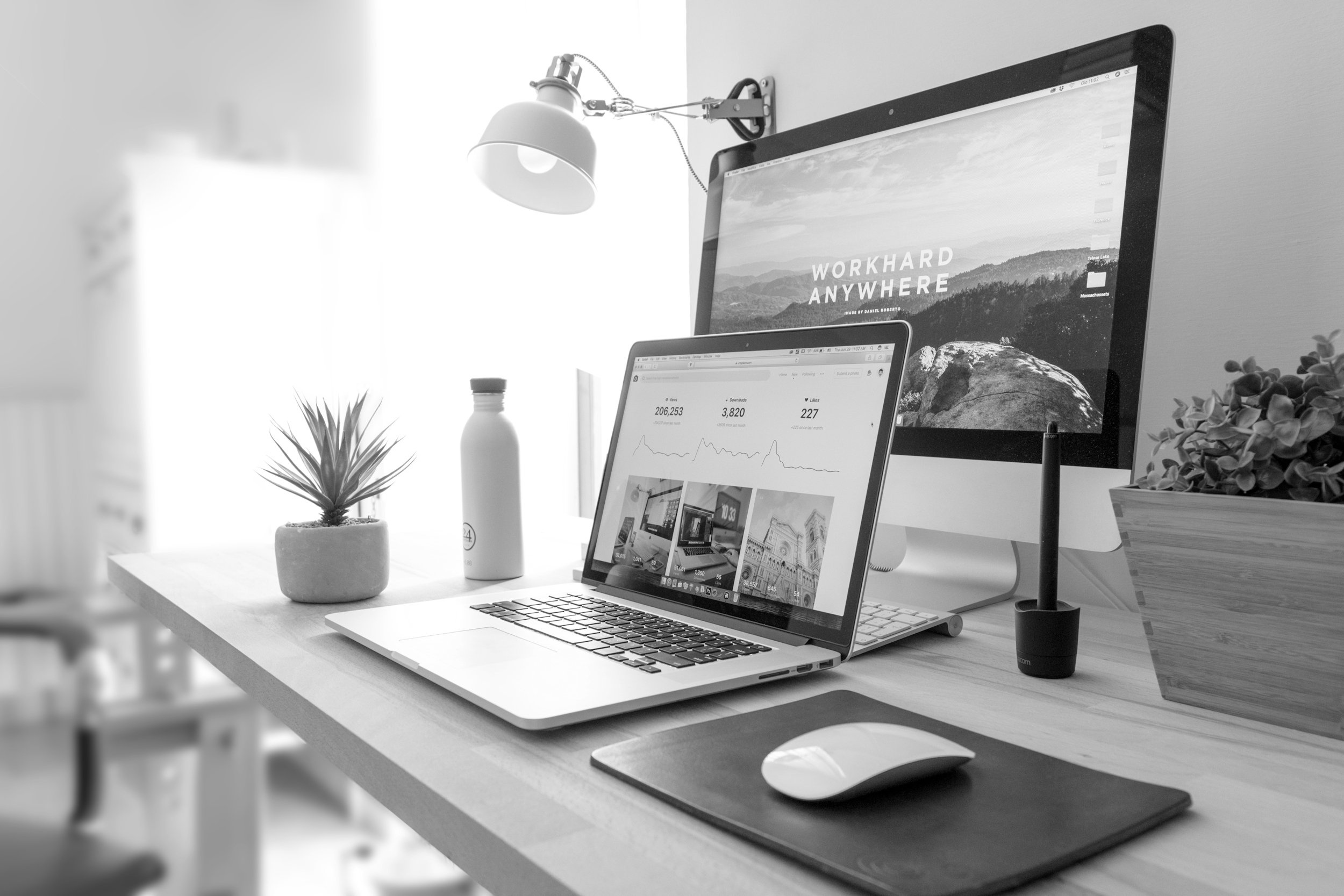 A modern workspace with a desktop computer, a laptop, a mouse on a mouse pad, a potted plant, a water bottle, a wall-mounted lamp, and a large monitor displaying nature images and motivational text, all arranged on a wooden desk.