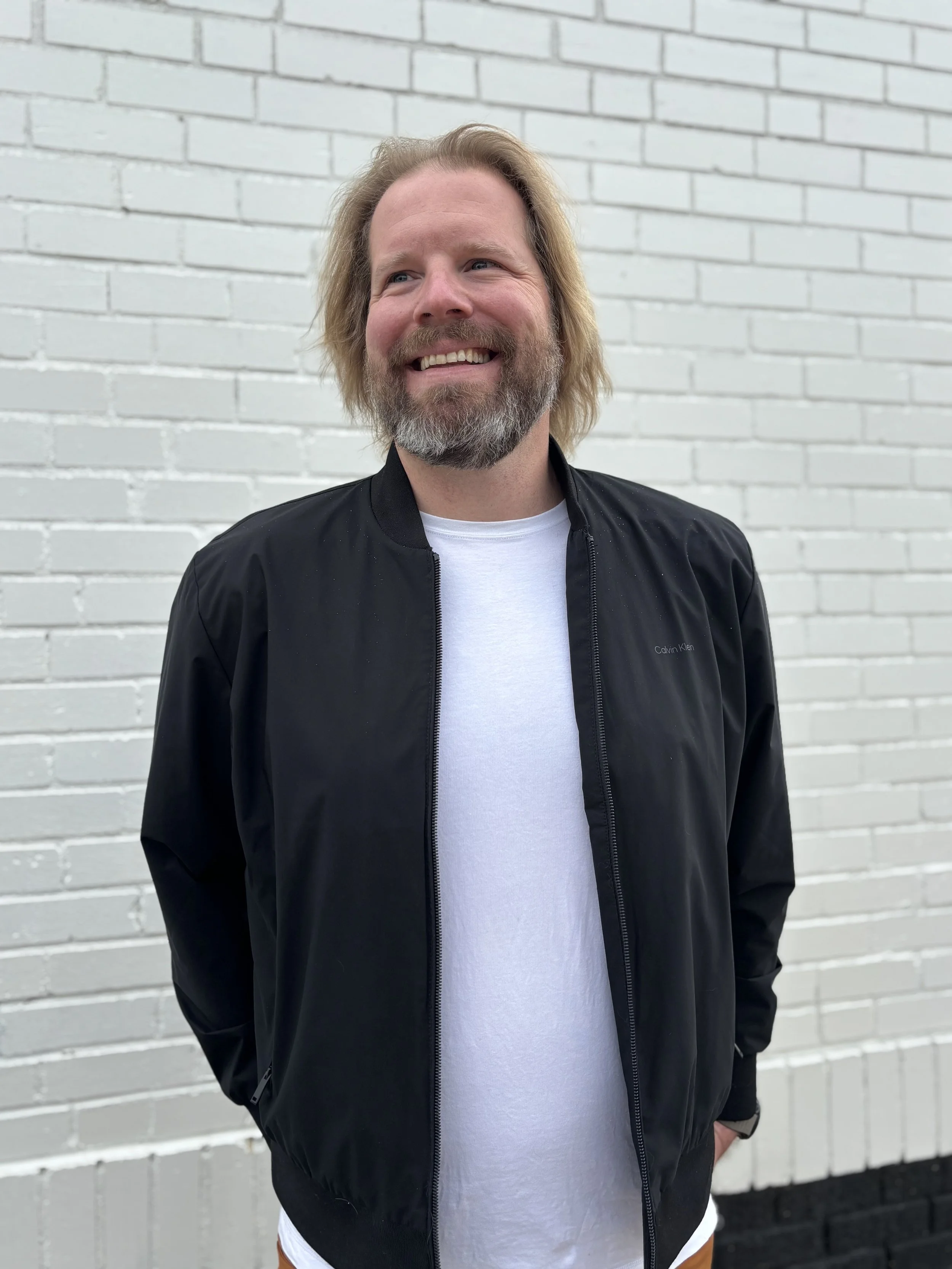 A man with shoulder-length blonde hair and a beard smiling, standing in front of a white brick wall. He is wearing a black jacket over a white T-shirt.