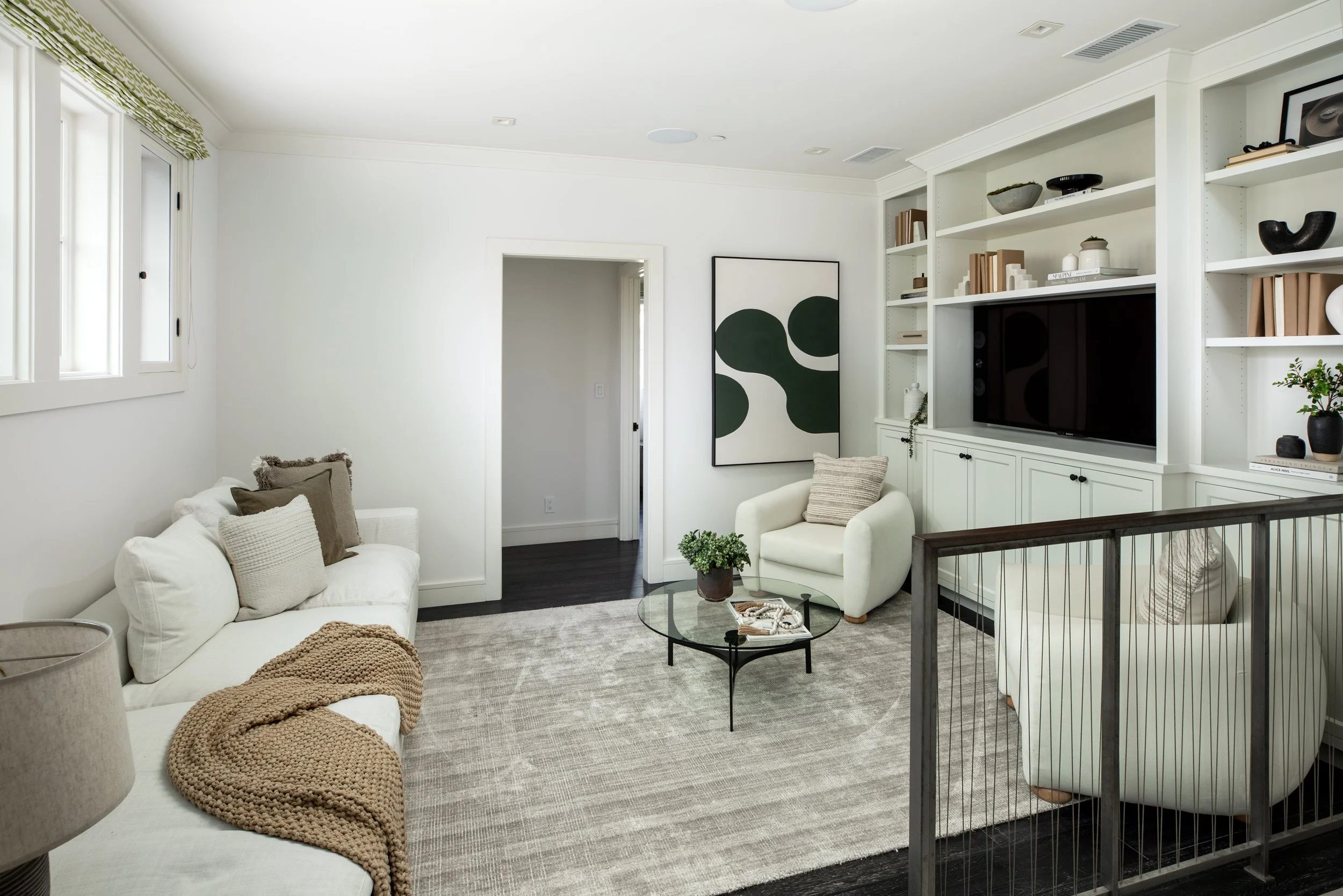 Living room with white sofa, beige knit throw, pillows, white armchair, glass coffee table with plant, built-in white shelves with decor, TV, artwork, and window with green and white shades.