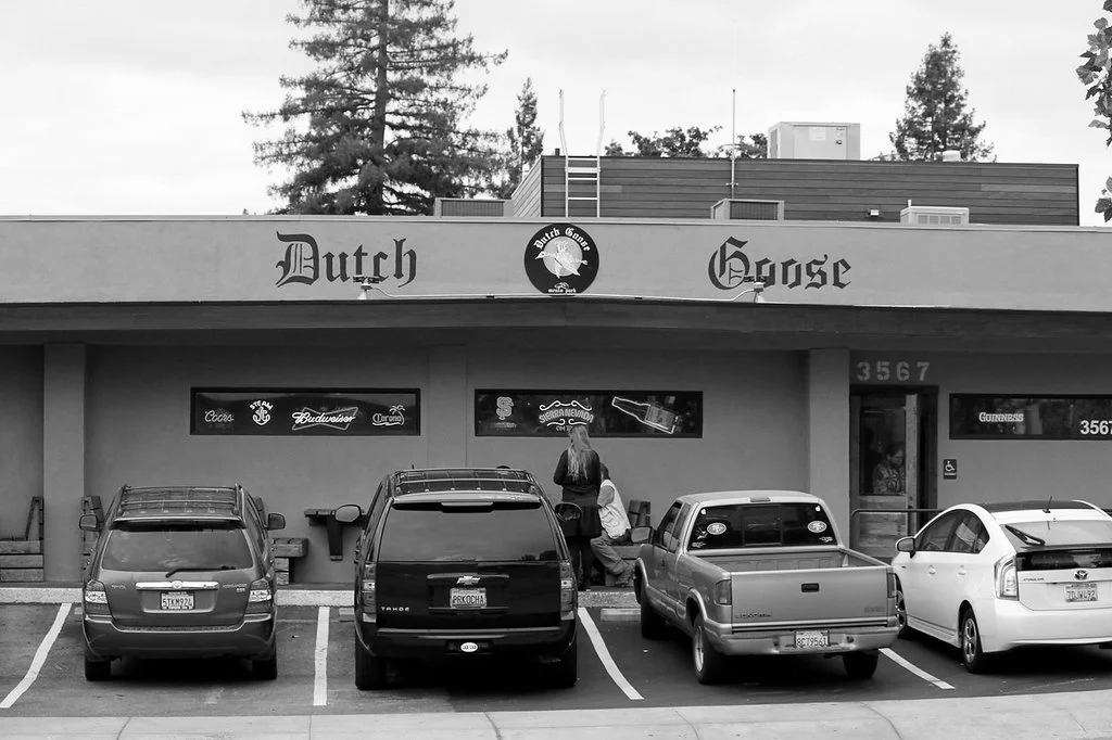 Front of a bar called Dutch Goose with parked cars, two people sitting on the sidewalk outside, and neon signs for various beer brands.