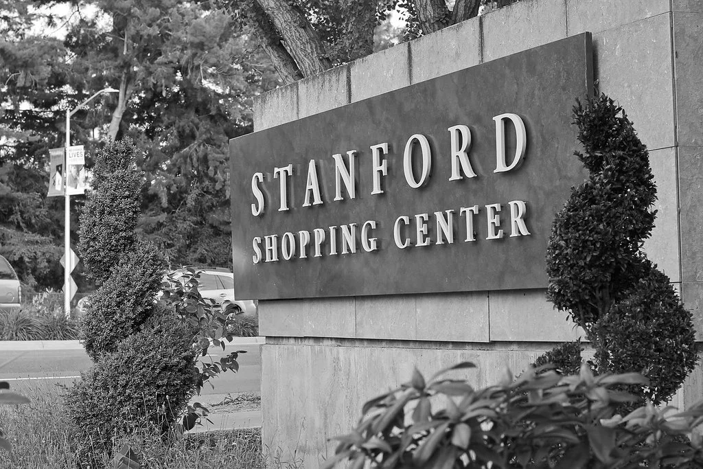 Black and white photo of a sign that reads 'Stanford Shopping Center,' surrounded by bushes and trees, near a road with vehicles and a street sign in the background.