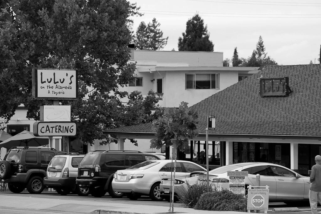 A parking lot in front of Lulu's on the Alameda restaurant, with several parked cars and a catering sign, surrounded by trees and buildings.