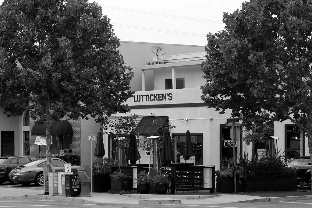 Black and white photo of a small shopping plaza featuring a business named Lutticken's with outdoor patio seating, trees, and parked cars.