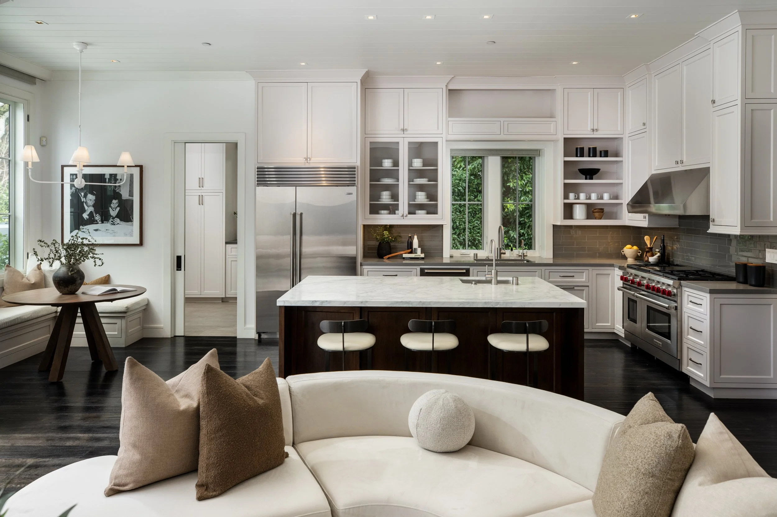 Modern open-concept kitchen with white cabinets, stainless steel appliances, an island with a marble top, and a white curved sofa with beige and brown pillows in the foreground.