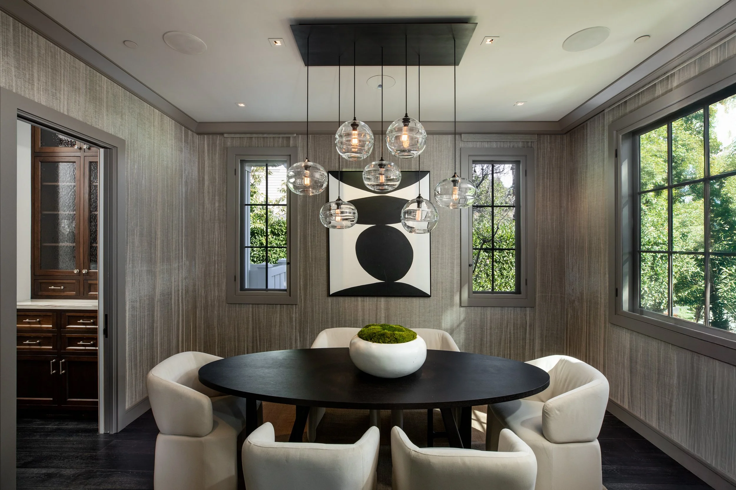 A modern dining room with a dark oval table, surrounded by six cream-colored chairs, with a bowl of moss in the center. Above, a cluster of glass pendant lights hangs from the ceiling. The room has two windows with grey frames, and a large black and 