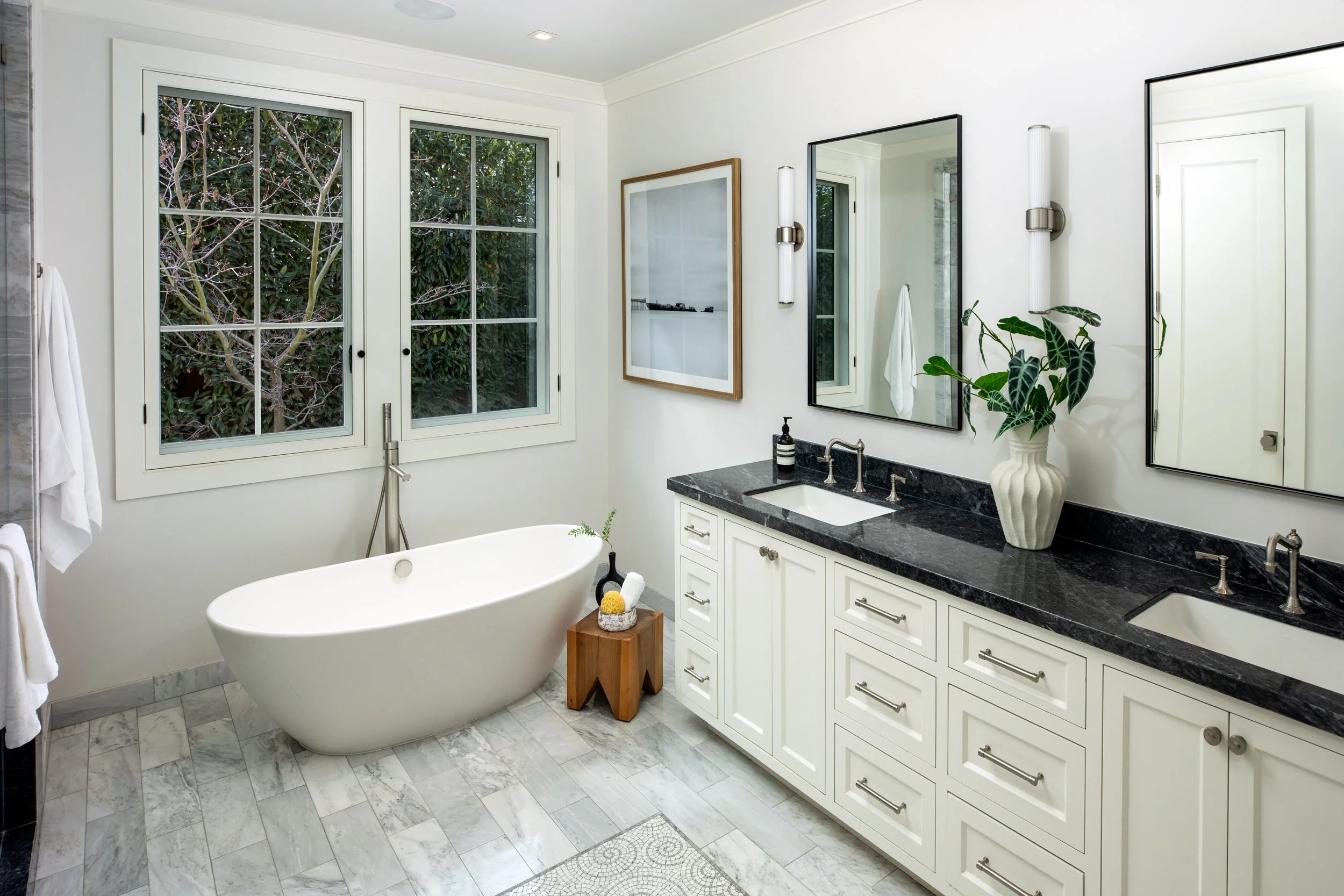 Modern bathroom with white walls, double sinks with black countertop, large mirror, bathtub, wooden side table, and window with greenery outside.
