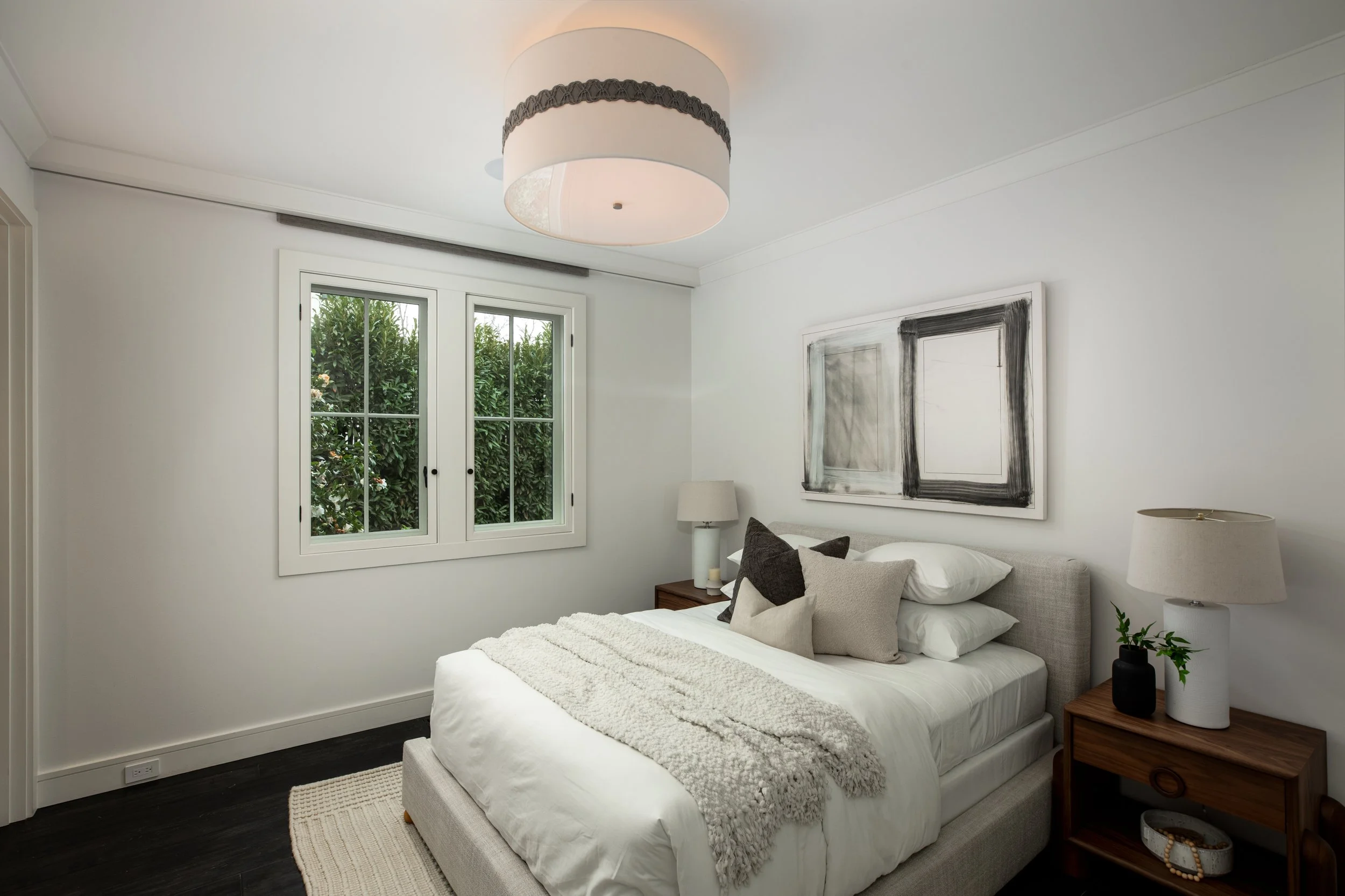 A modern bedroom with white walls and a bed with multiple pillows, a throw blanket, and a window showing greenery outside. There are two bedside tables with lamps and a simple abstract painting above the bed.