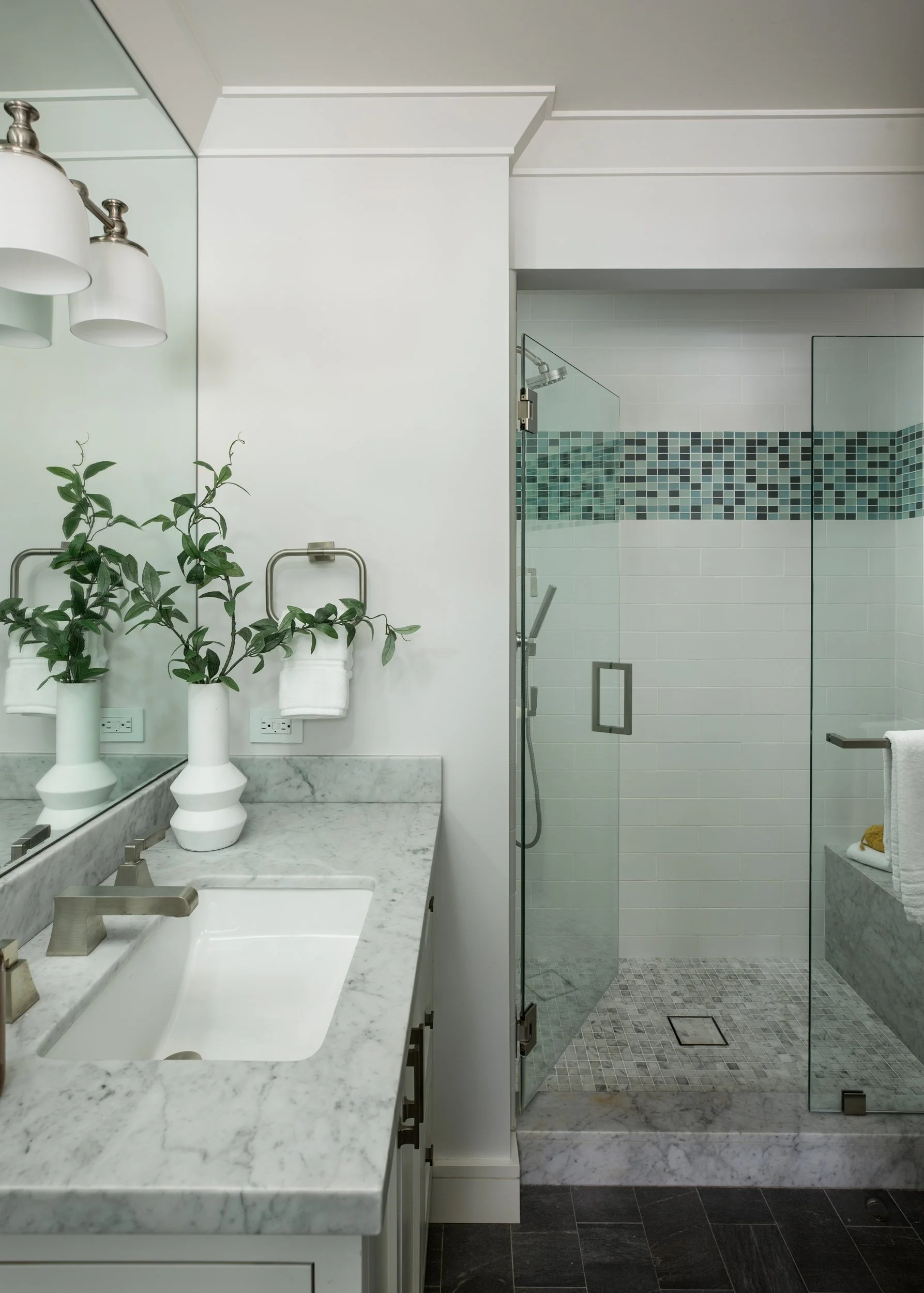 Modern bathroom with white countertop, sink, and two decorative vases with greenery. Glass shower enclosure with tiled shower area featuring a strip of colorful mosaic tiles.