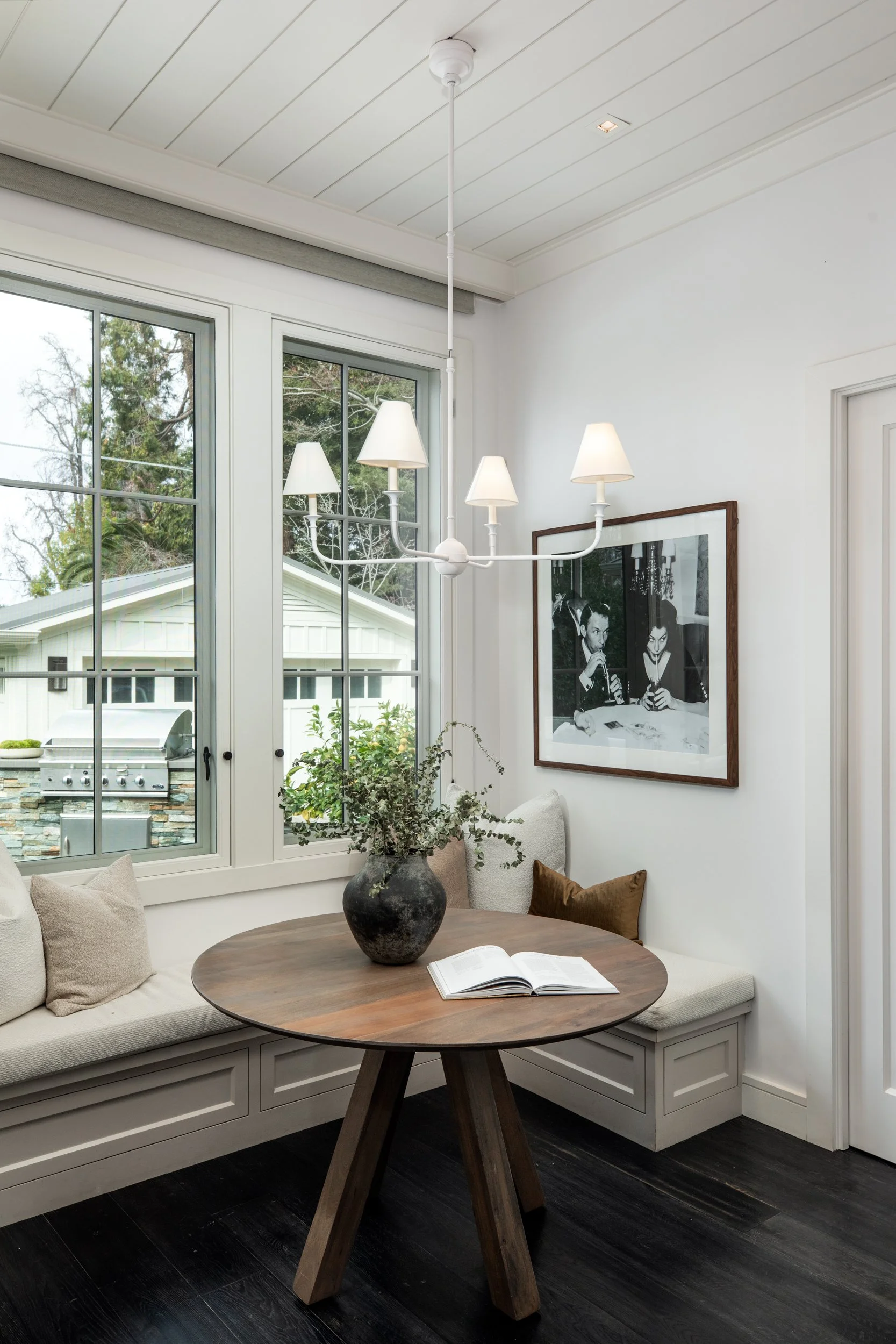 Interior of a cozy nook with a round wooden table and cushioned bench, black and white artwork on white walls, large windows revealing a garden, and a modern white chandelier with lampshades.