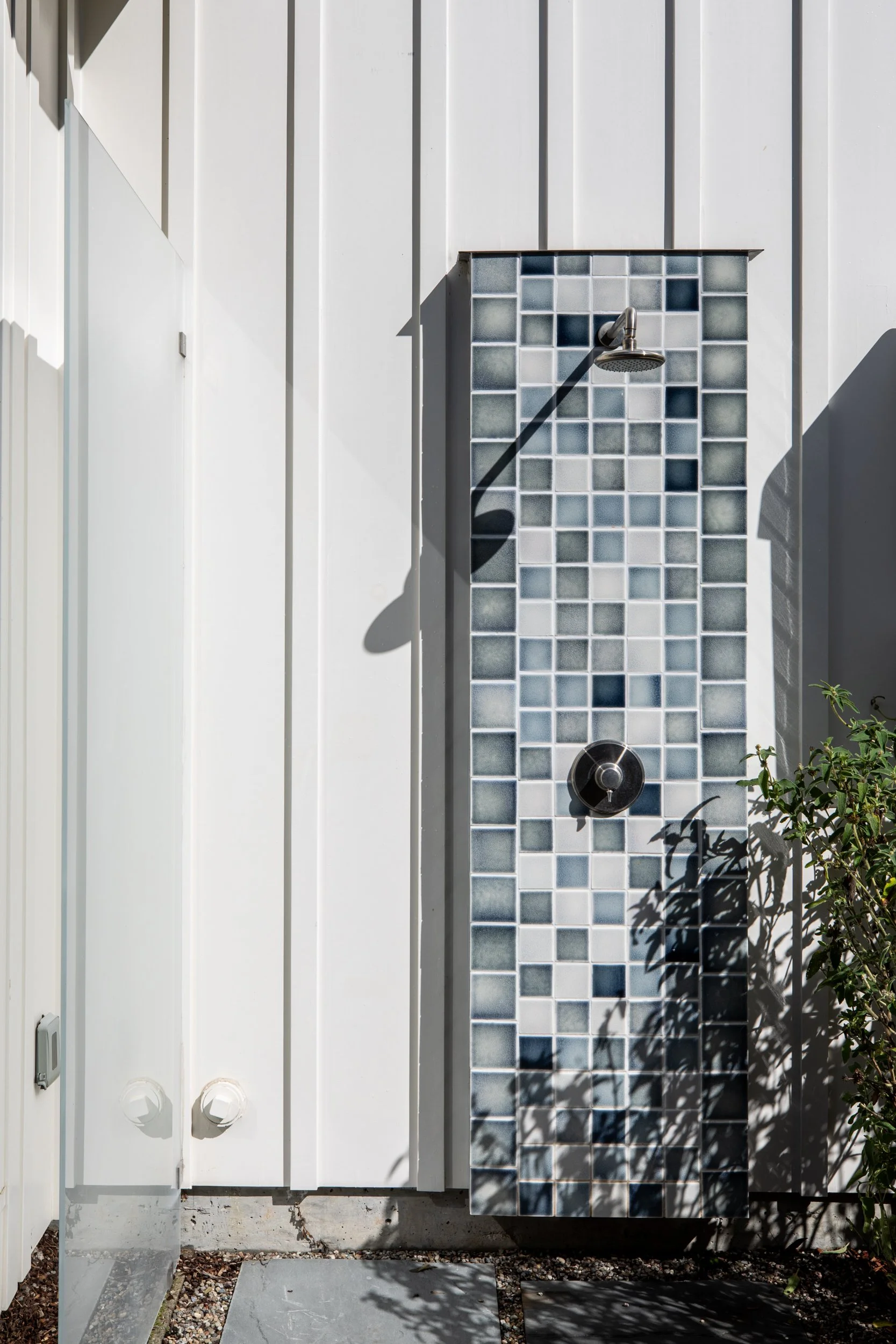 Outdoor shower with gray and black tiles on a wooden panel, mounted on a white exterior wall, with sunlight and shadow.