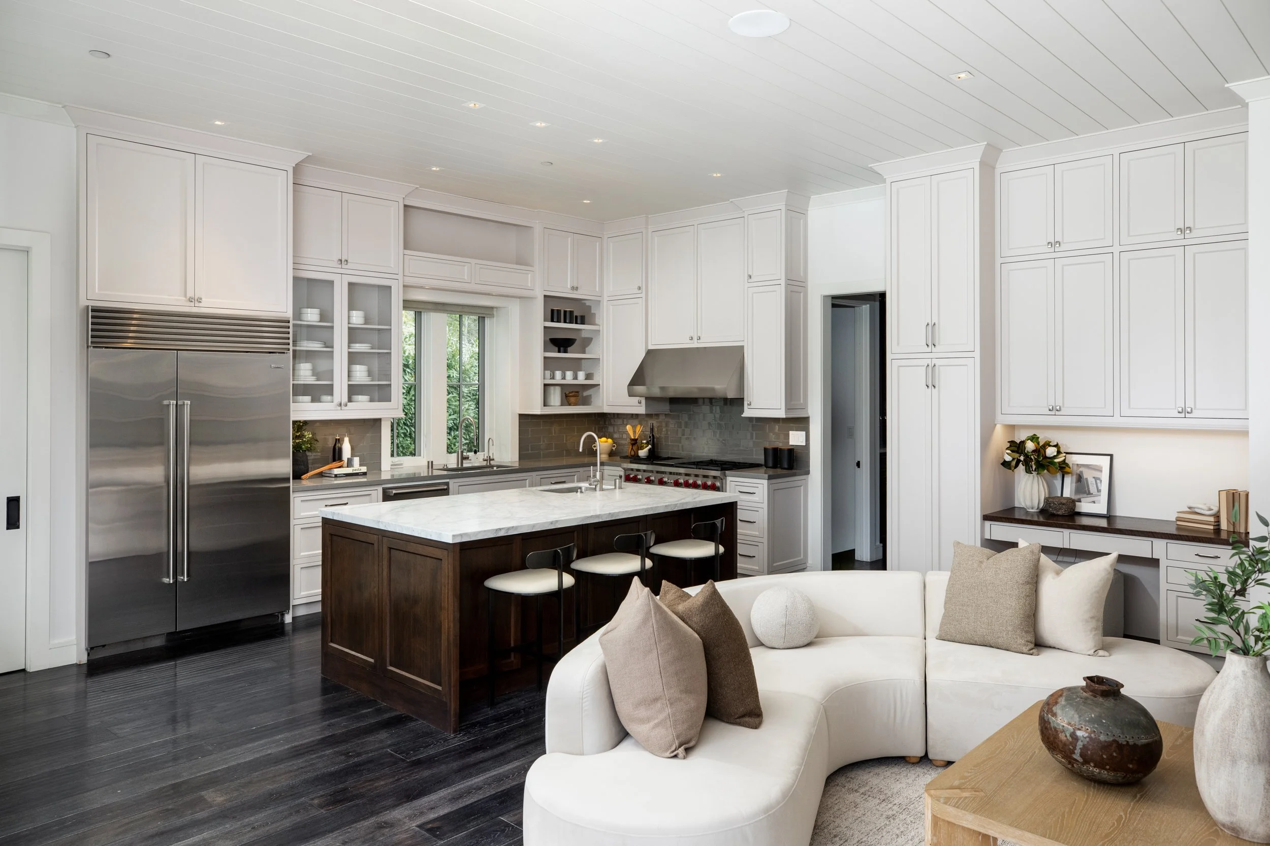 Modern kitchen with white cabinets, stainless steel refrigerator, island with marble countertop, dark hardwood floors, and a white sofa with beige and brown pillows in the living area.