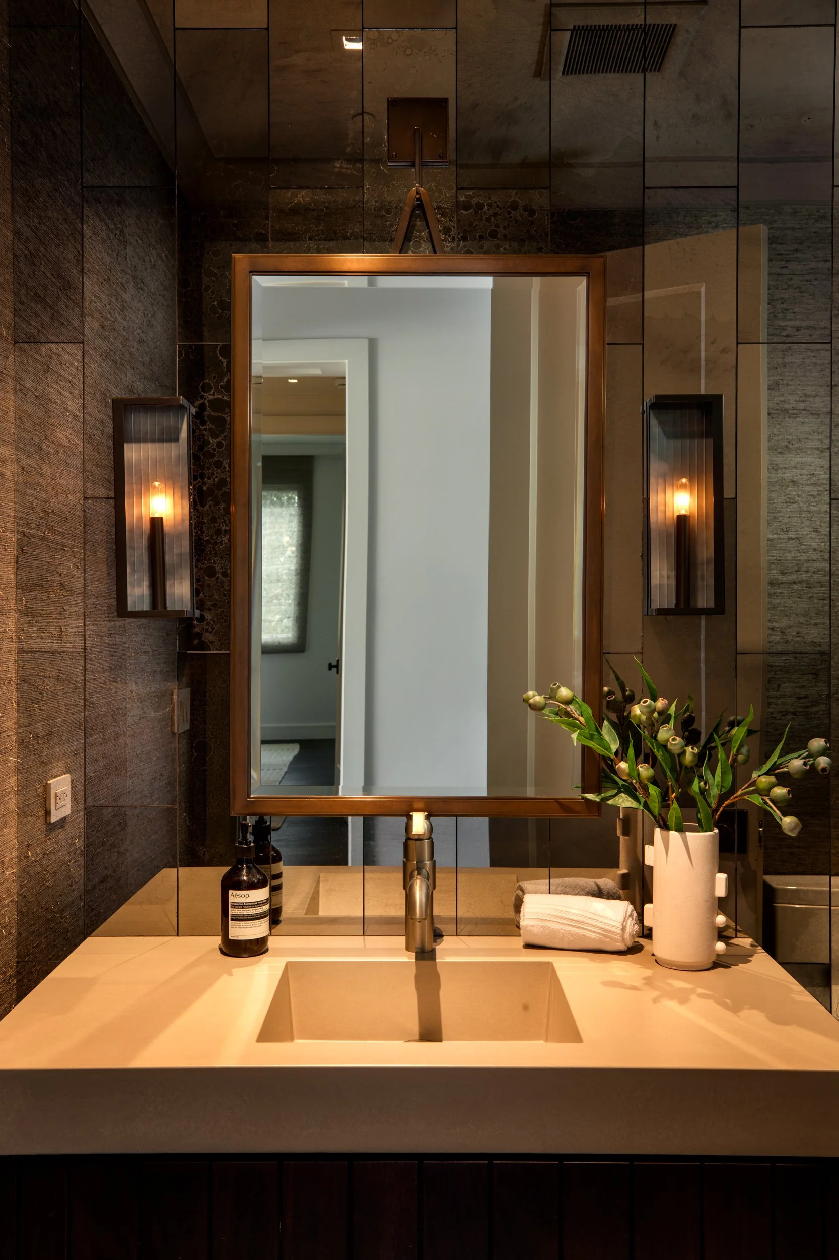 Bathroom vanity area with a large mirror, a sink, a soap dispenser, folded towels, and a vase with green foliage, with reflection showing another room and window.