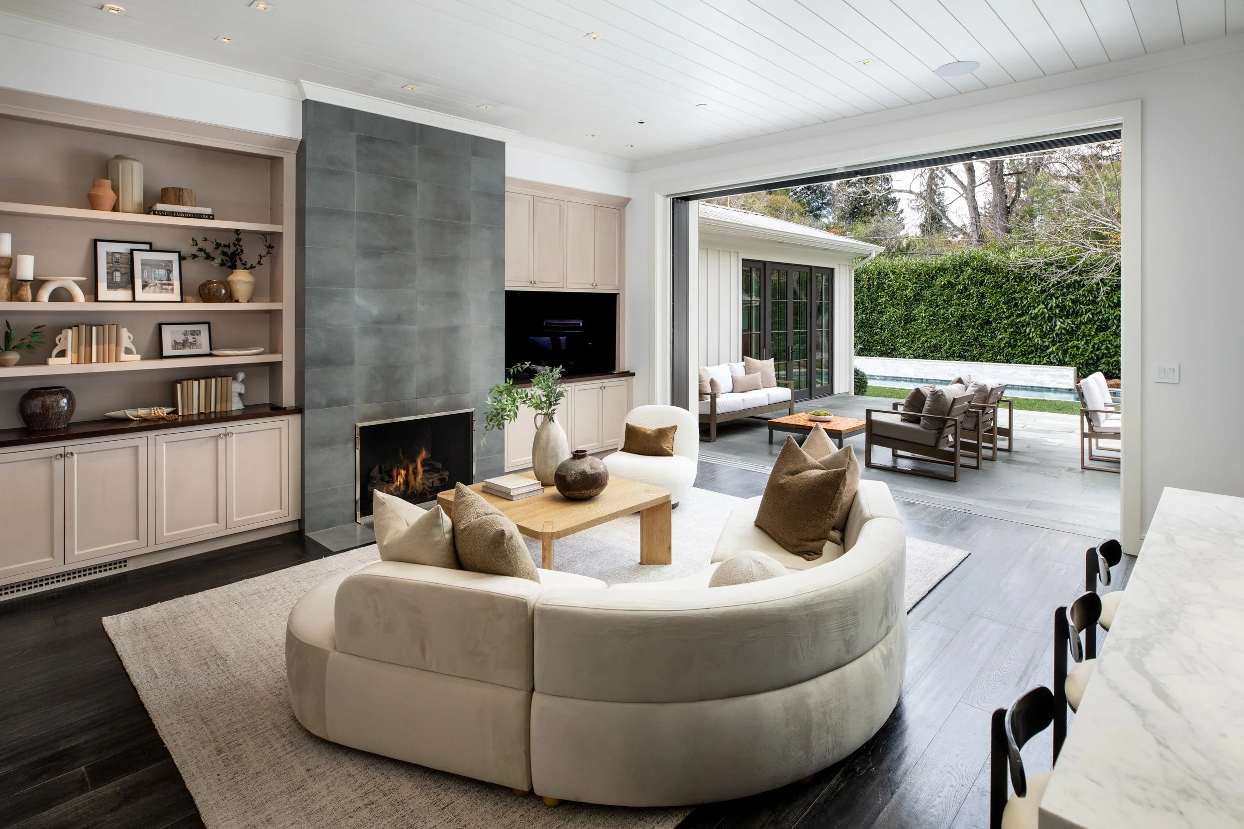 Bright living room with a white curved sofa, wooden coffee table, and a fireplace with a gray tiled surround. Large sliding doors open to an outdoor patio with seating and a backyard with a pool and greenery.