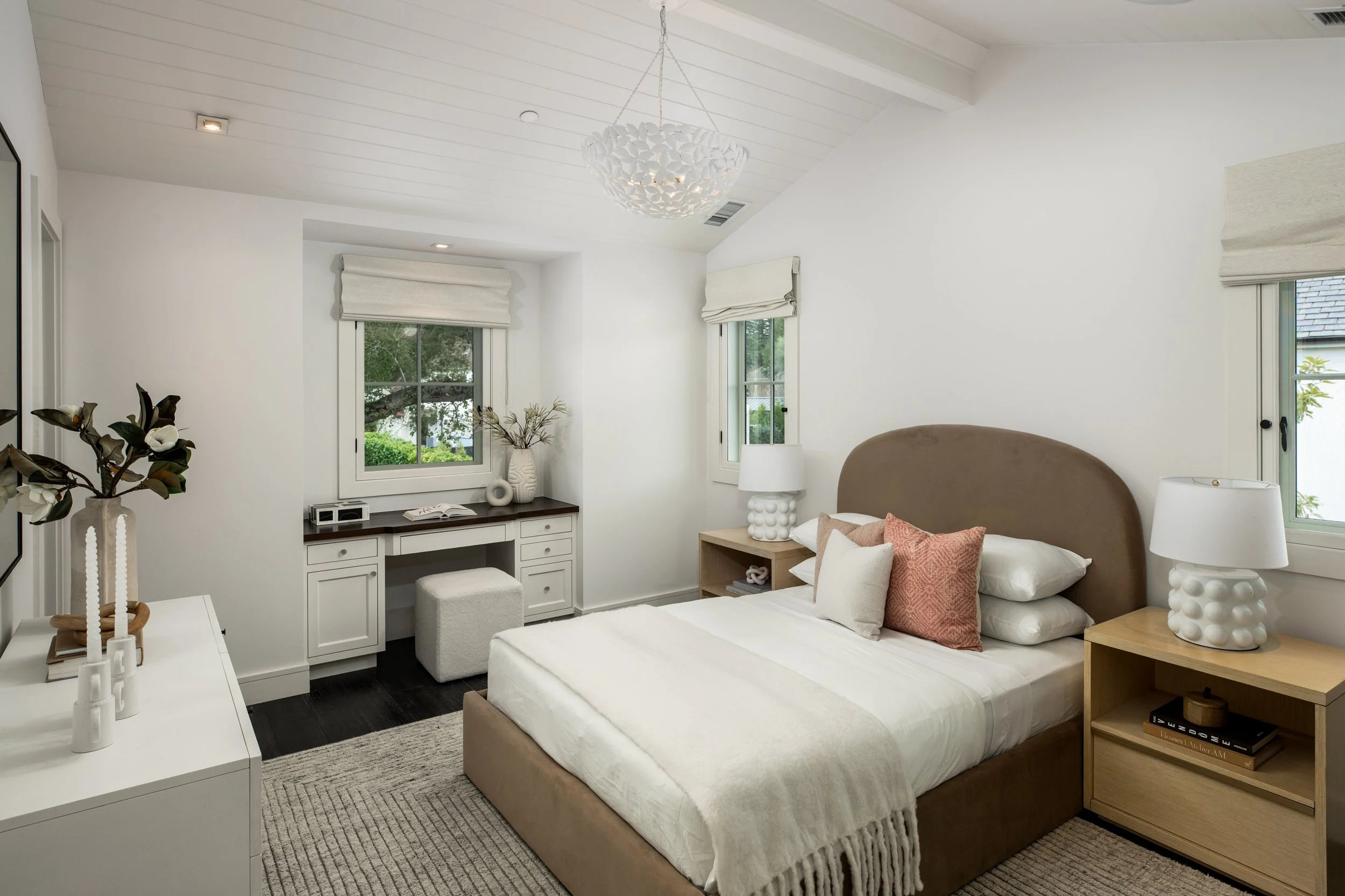 A bedroom with white walls, a beige bed with pillows, two wooden nightstands with lamps, a window with blinds, and a built-in desk underneath a window.