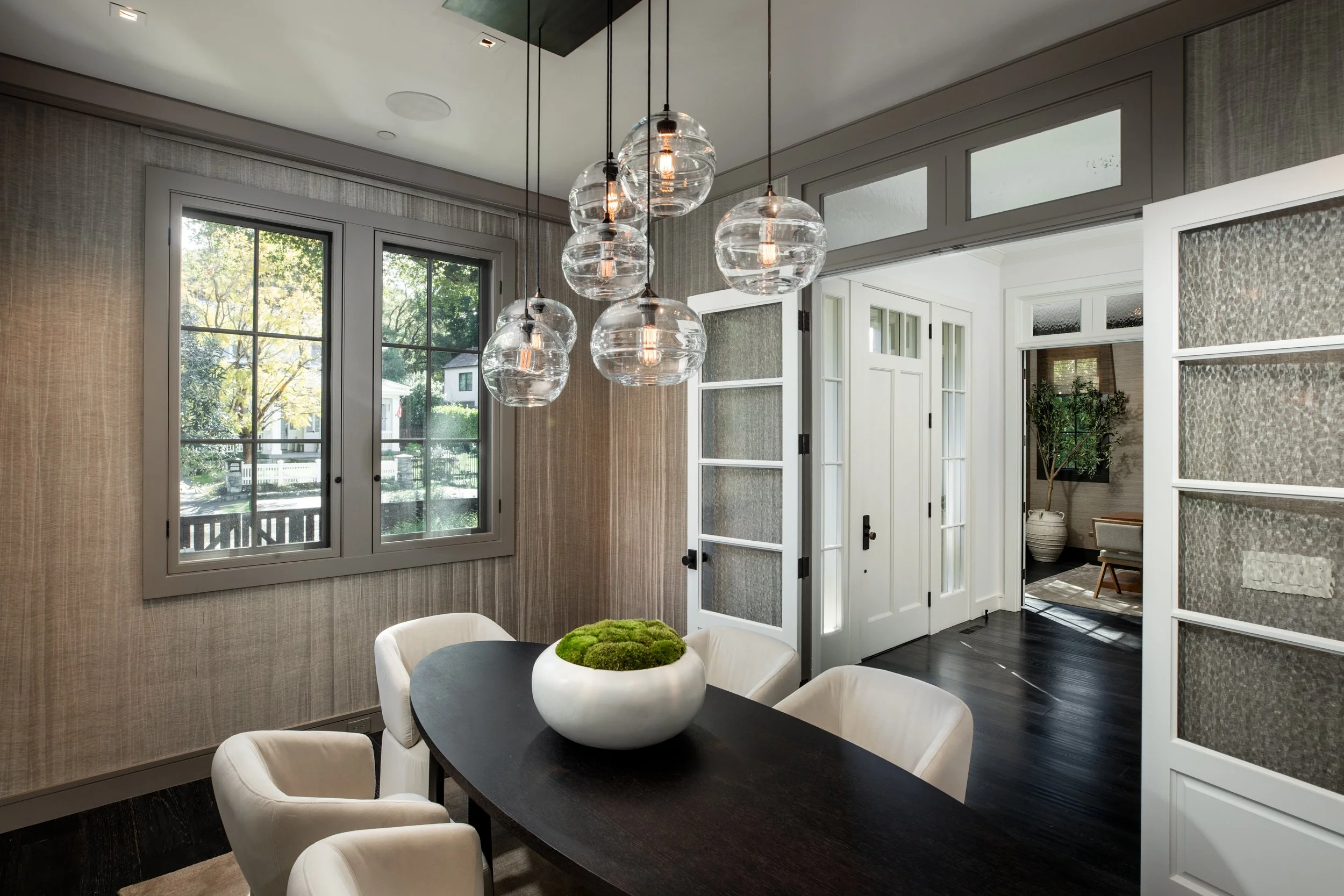Modern dining room with a dark oval table, six white chairs, a white bowl of green moss, large glass pendant lights, windows showing a yard, and open doors leading to another room.