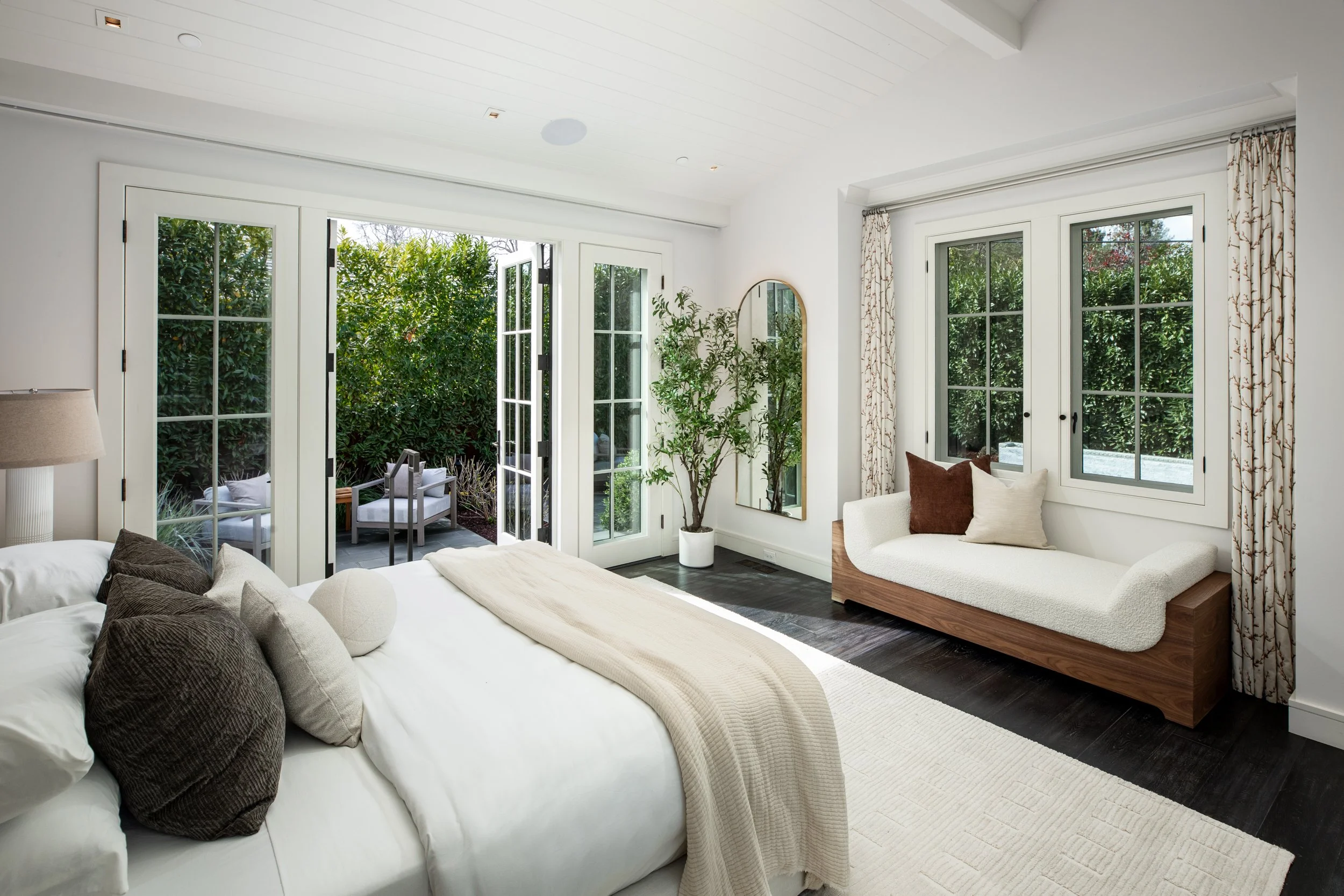 Bright bedroom with white walls, large windows, and a balcony door looking to a green outdoor area. The bed has white bedding and brown and beige throw pillows. There's a cushioned window seat with pillows, a potted plant, and a mirror on the wall.