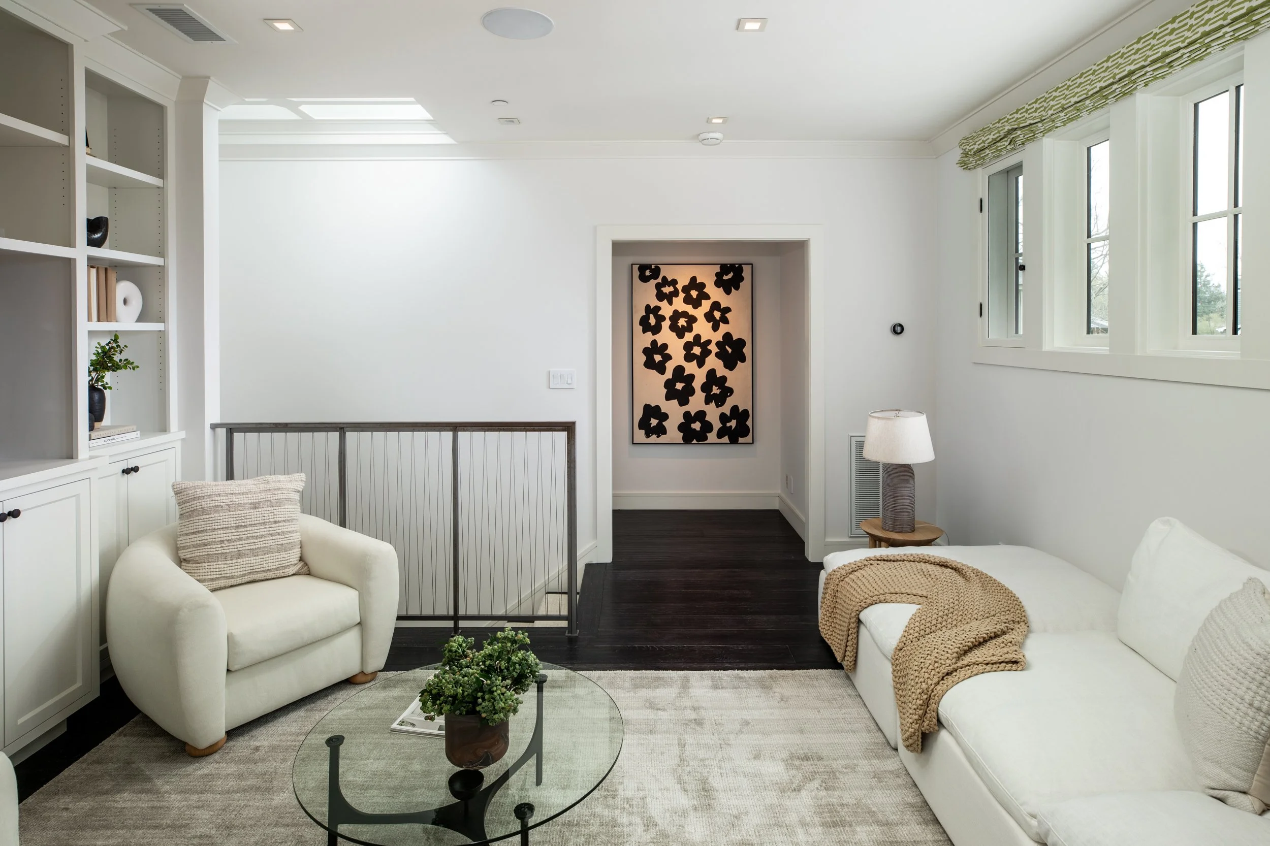 A bright living room with white walls, a white sofa with a beige throw blanket, a white armchair with a beige pillow, a glass coffee table with a potted plant, and windows with green valances. There's a black and white floral artwork on the back wall