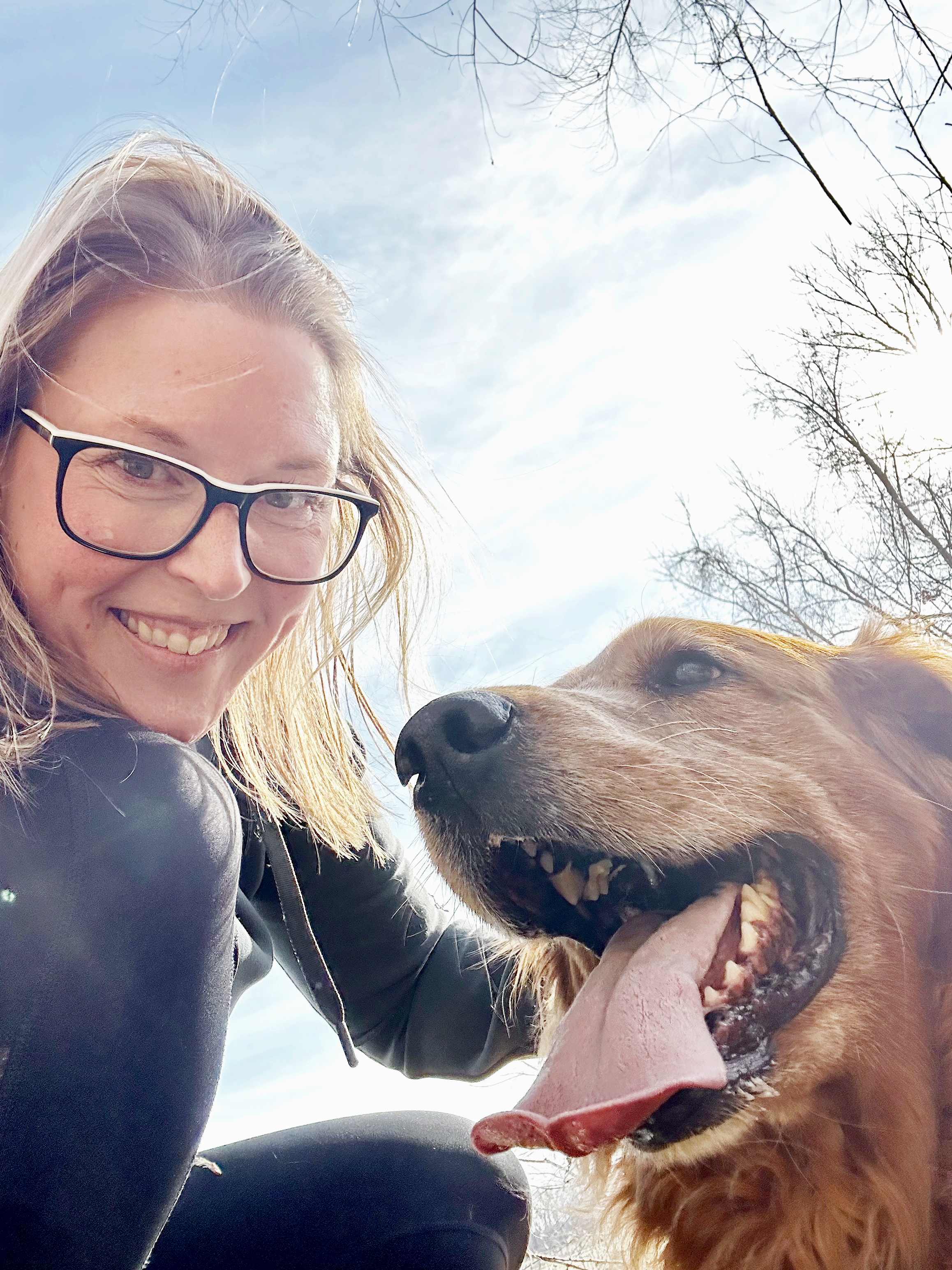 A woman with glasses and a black leather jacket smiling next to a golden retriever with its tongue out, outdoors with a cloudy sky and tree branches in the background.