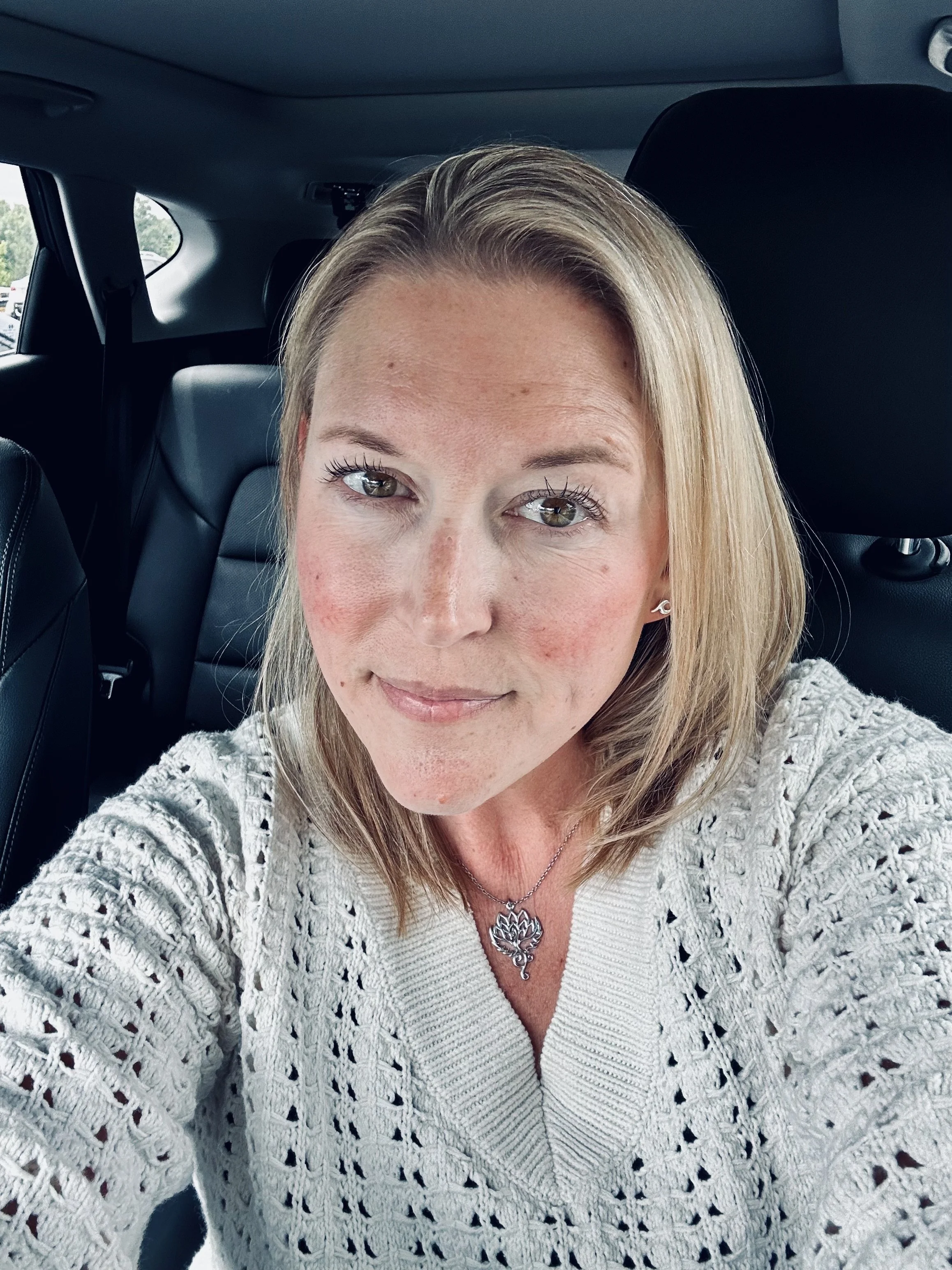 Close-up of a woman with blonde hair taking a selfie inside a car, wearing a cream-colored knitted sweater and a silver necklace with a lotus flower pendant.