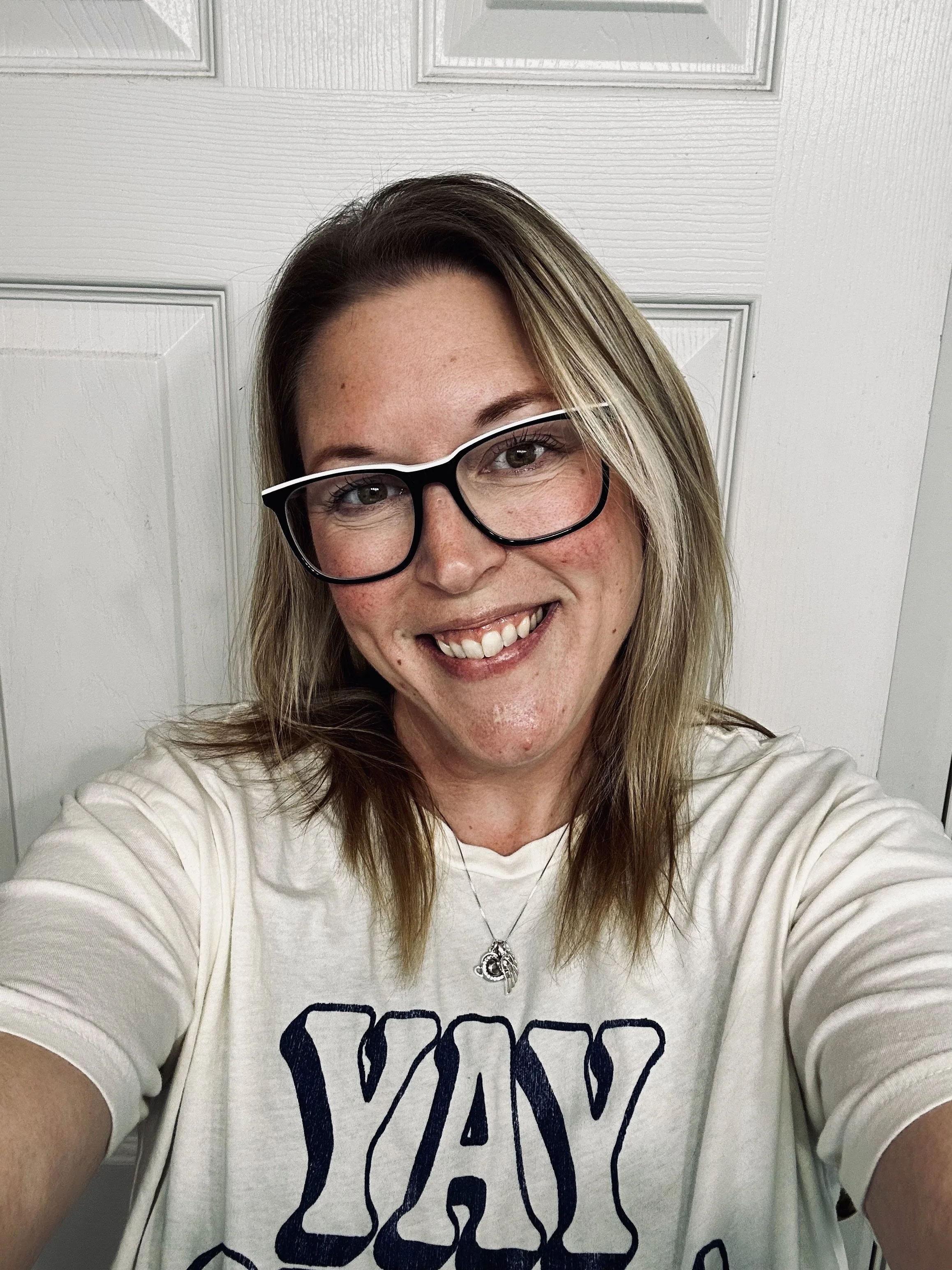 A woman with shoulder-length blonde hair, wearing glasses and a white T-shirt, smiling and taking a selfie in front of a white door.