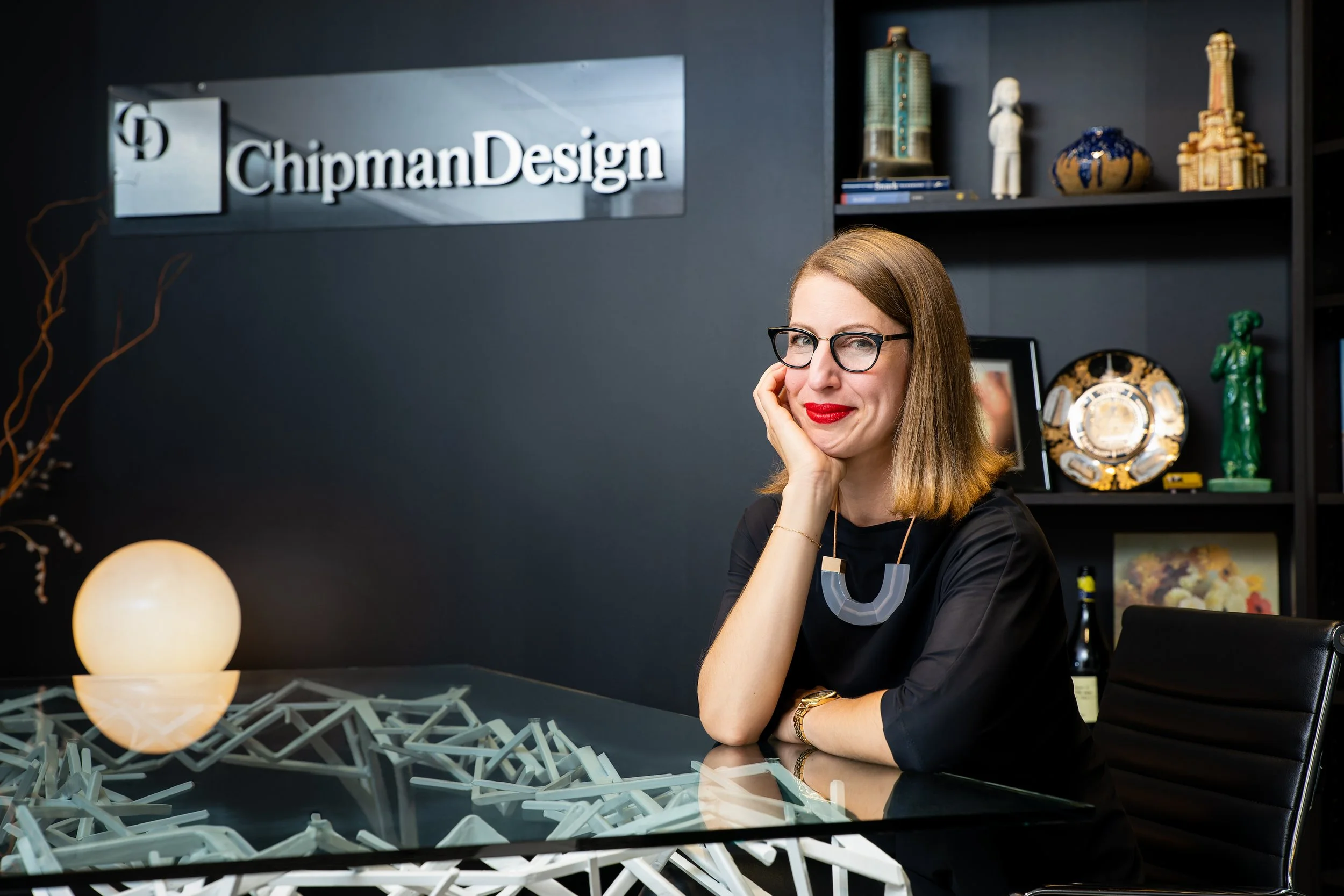 A woman with glasses, red lipstick, and shoulder-length light brown hair sitting at a desk with a decorative object made of light-colored pieces, in an office with a black wall that has a "ChipmanDesign" sign. There are shelves with art and decorative items in the background, and a glowing spherical light on the desk.
