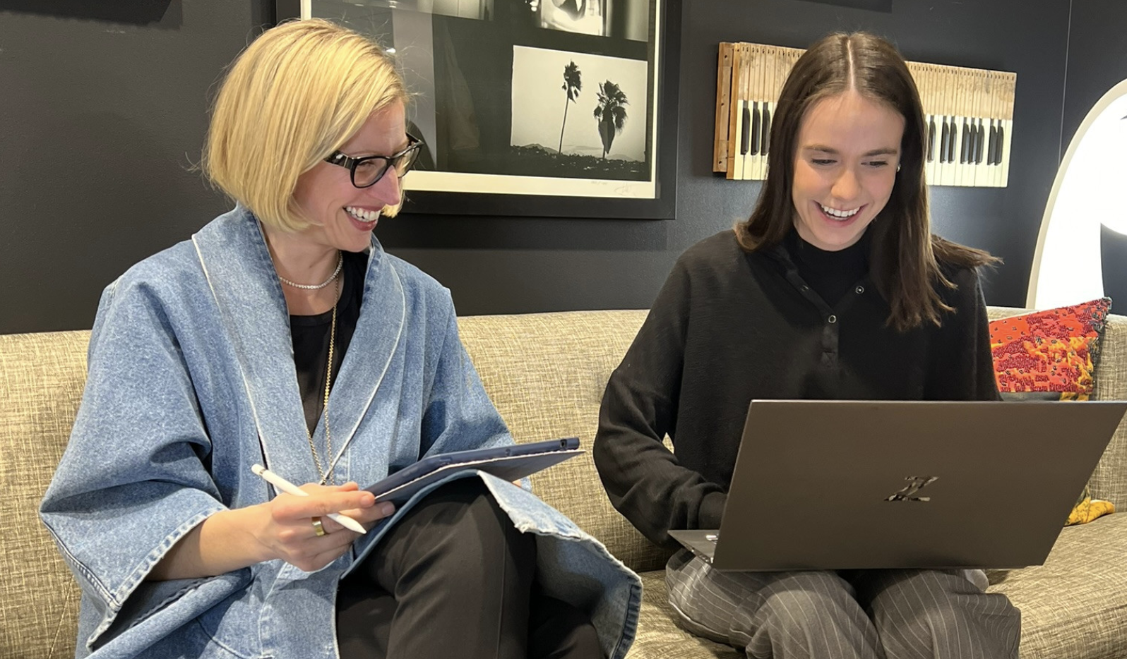 Two women sitting on a beige sofa, smiling and looking at a laptop and a notepad, in a modern room with black walls and wall art.