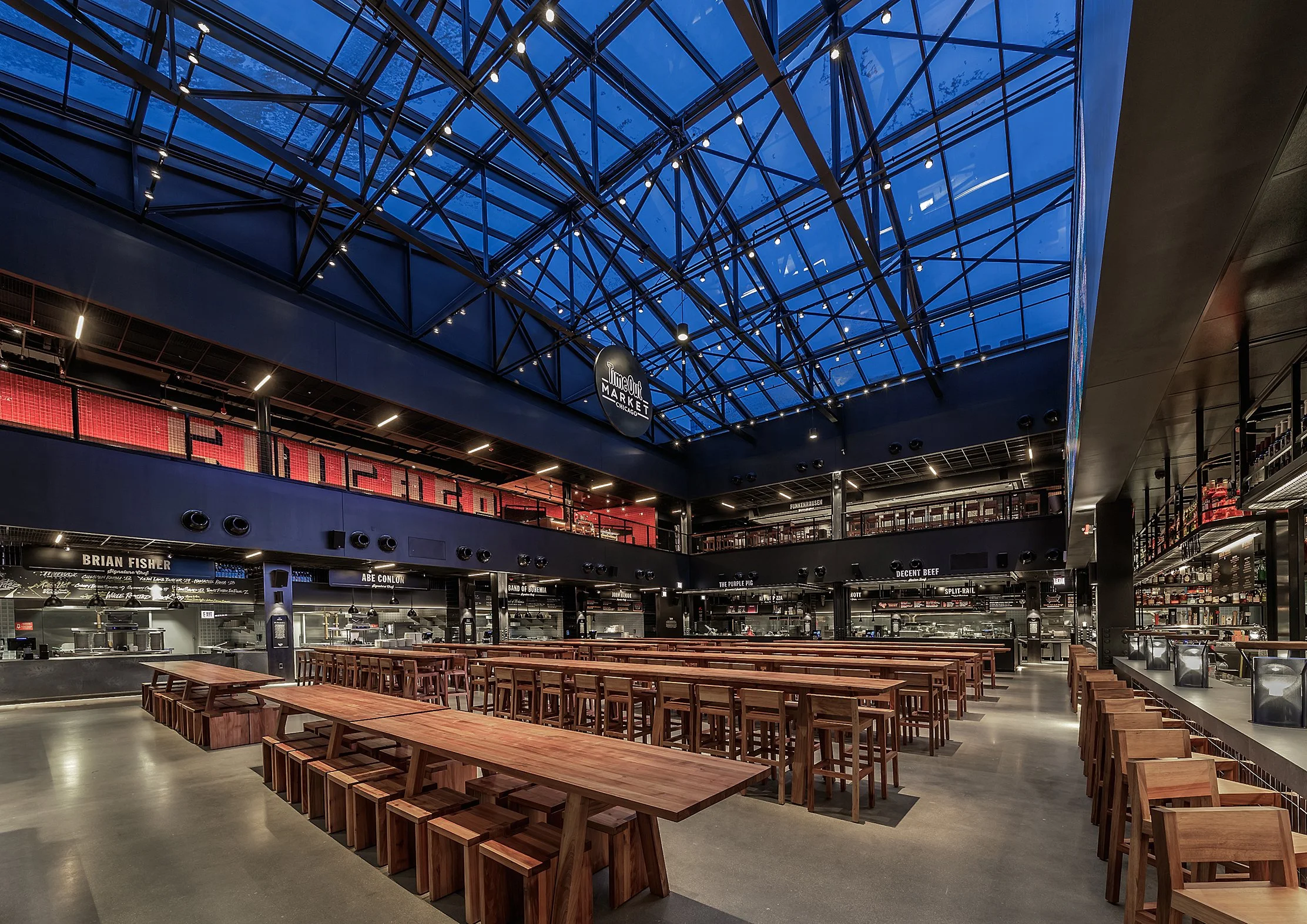Interior of a modern marketplace with wooden tables and benches, glass ceiling, and a sign that says 'Time Out Market Chicago'.