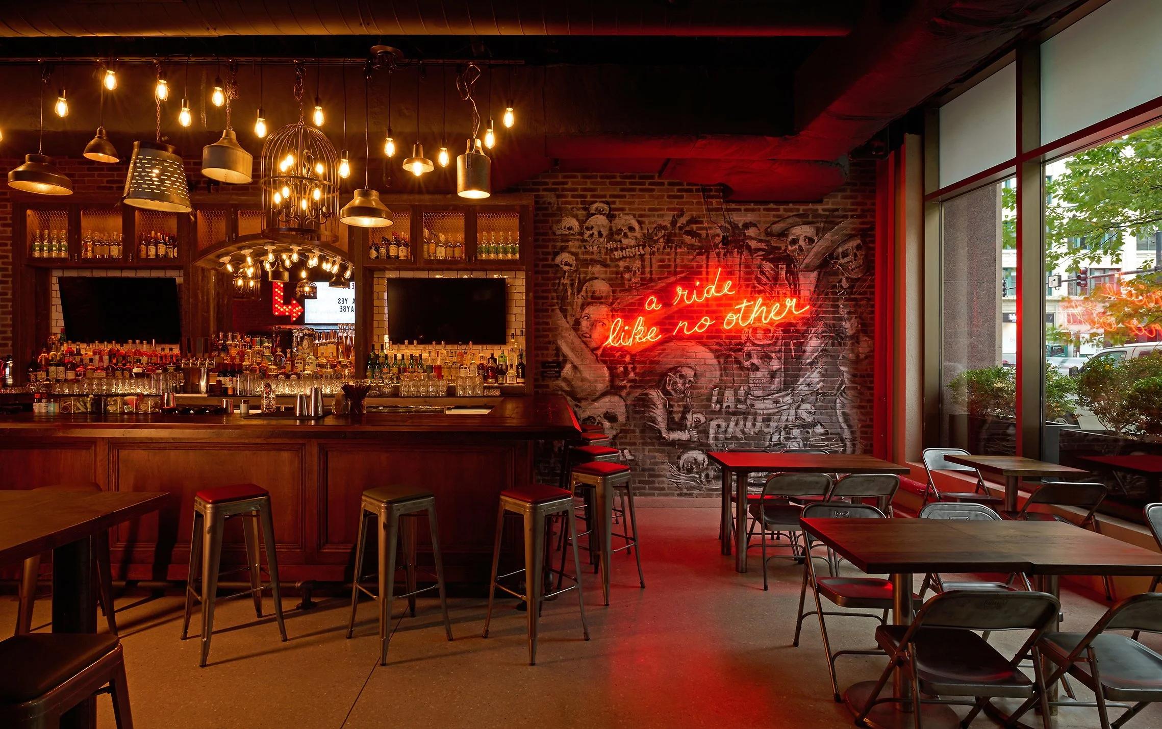 Interior of a dimly lit bar or restaurant with wooden tables and chairs, industrial-style lighting fixtures hanging from the ceiling, a brick wall with a mural of skulls, and a red neon sign that reads 'a ride like no other.' Large windows on the rig