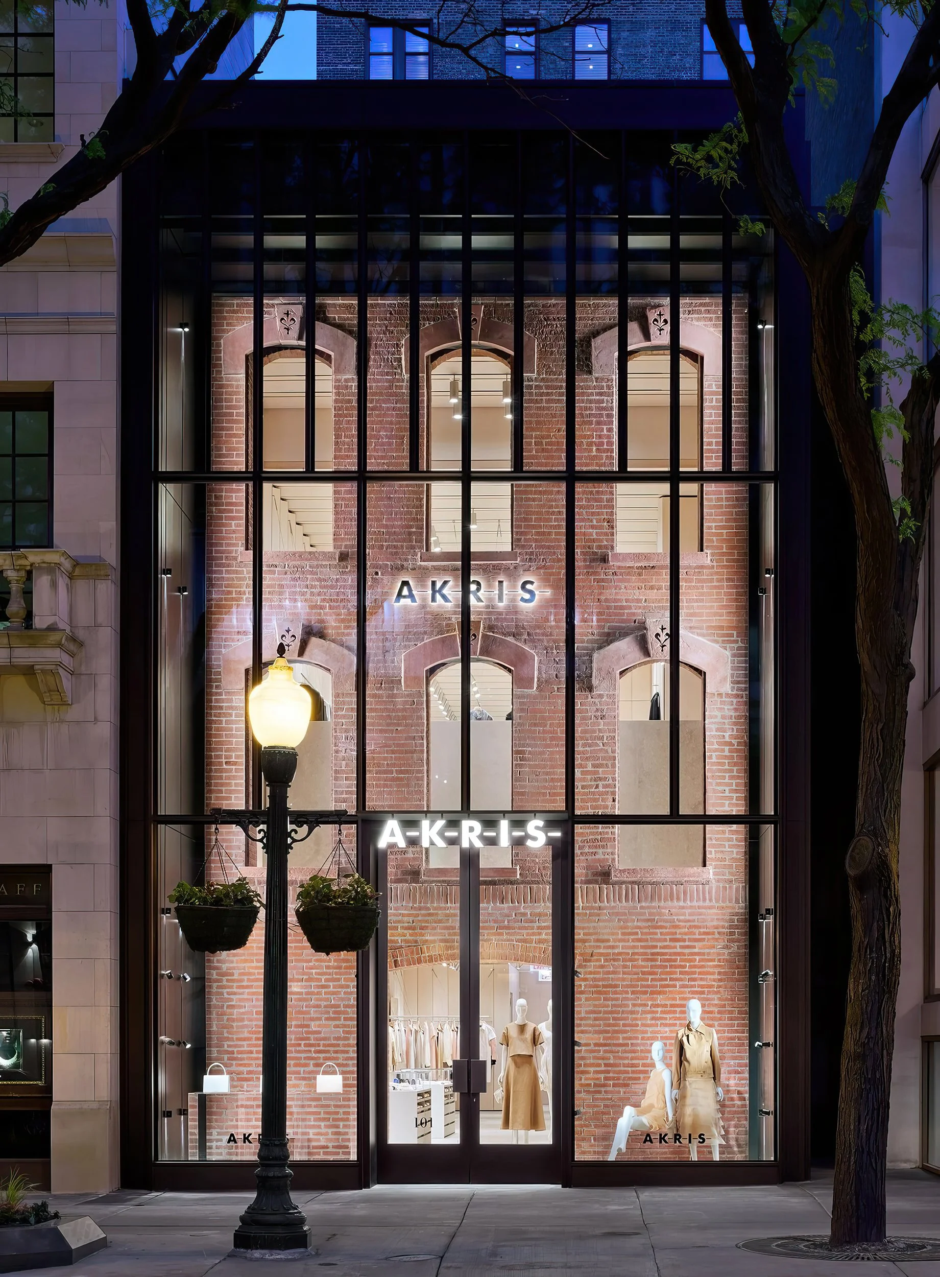 Storefront at night with large glass windows, displaying mannequins in beige clothing, and a sign that reads 'AKRIS' in illuminated letters.
