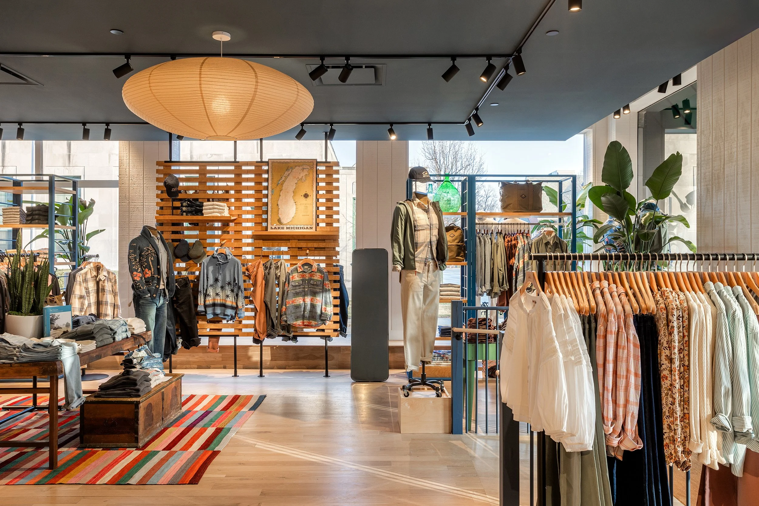 Interior of a clothing store displaying men's and women's apparel, shelves with folded clothes, racks of shirts and jackets, decorative plants, and a large paper lantern ceiling light. Large windows let in natural light.