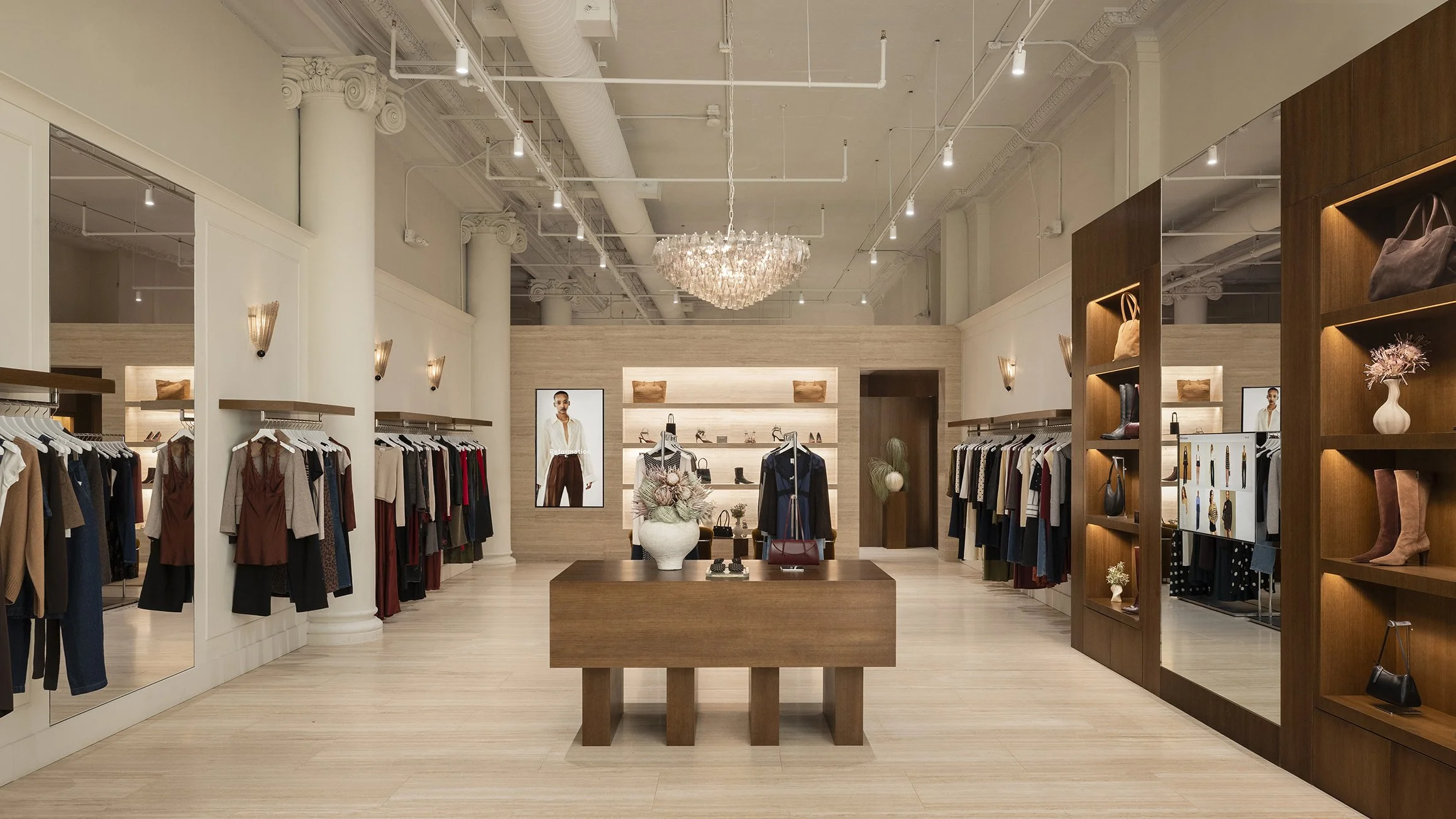 Interior of a clothing store with racks of clothing, decorative shelves with handbags and accessories, a chandelier hanging from the ceiling, and framed fashion posters on the wall.