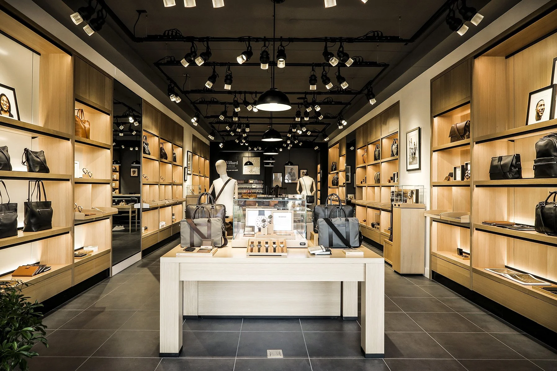 Interior of a retail store displaying handbags, accessories, and jewelry on wooden shelves and tables, with mannequins showcasing items, and framed pictures on the walls, lit by ceiling lamps.