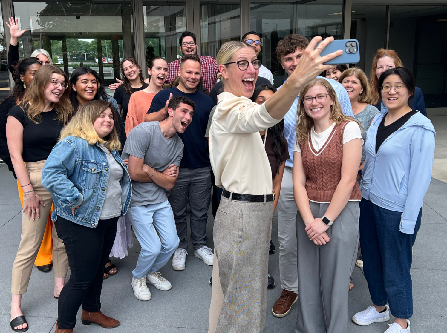 A group of diverse people gathered outside, taking a selfie together with a woman holding a smartphone, all smiling and appearing joyful.