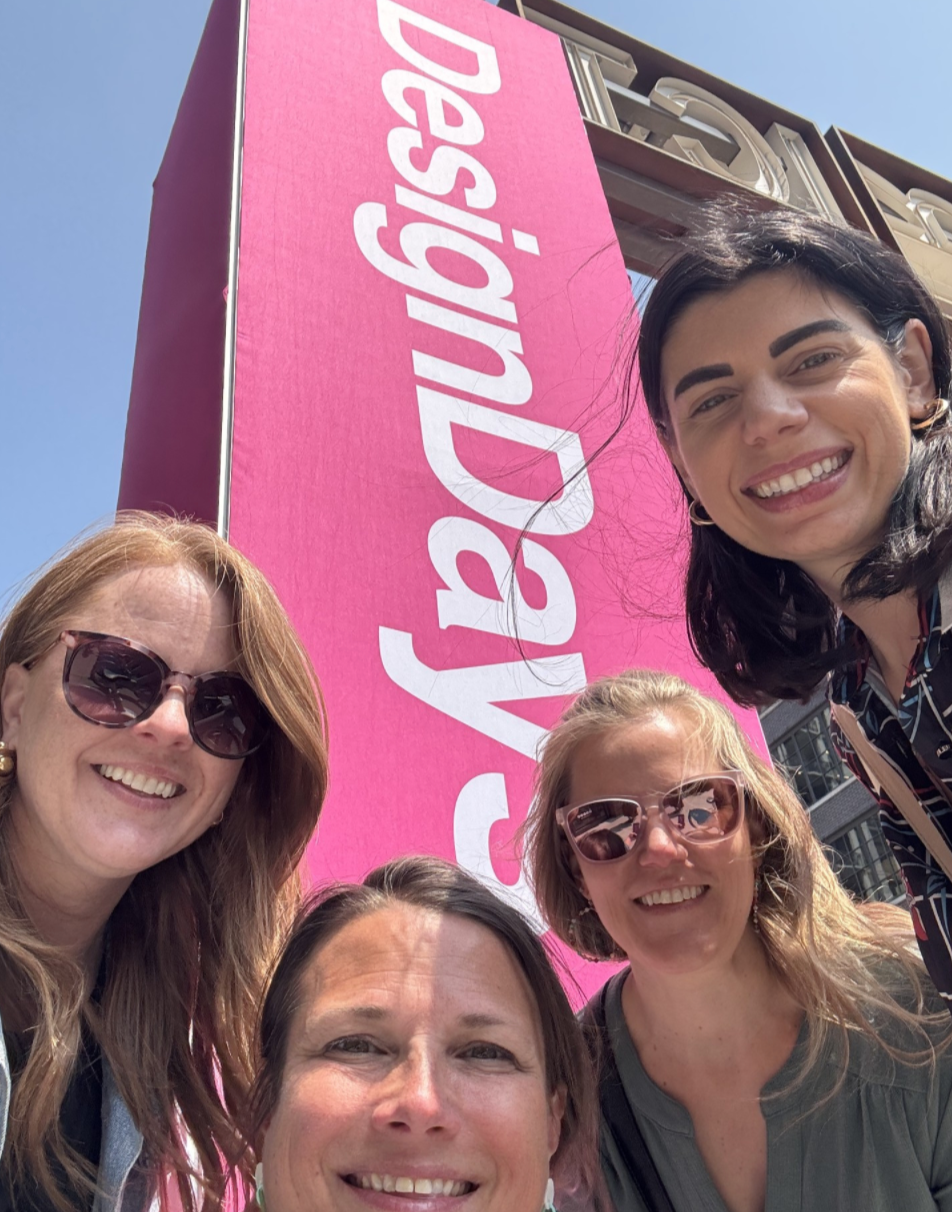 Group of five women smiling for a selfie in front of a large pink banner that says 'Desigual'.