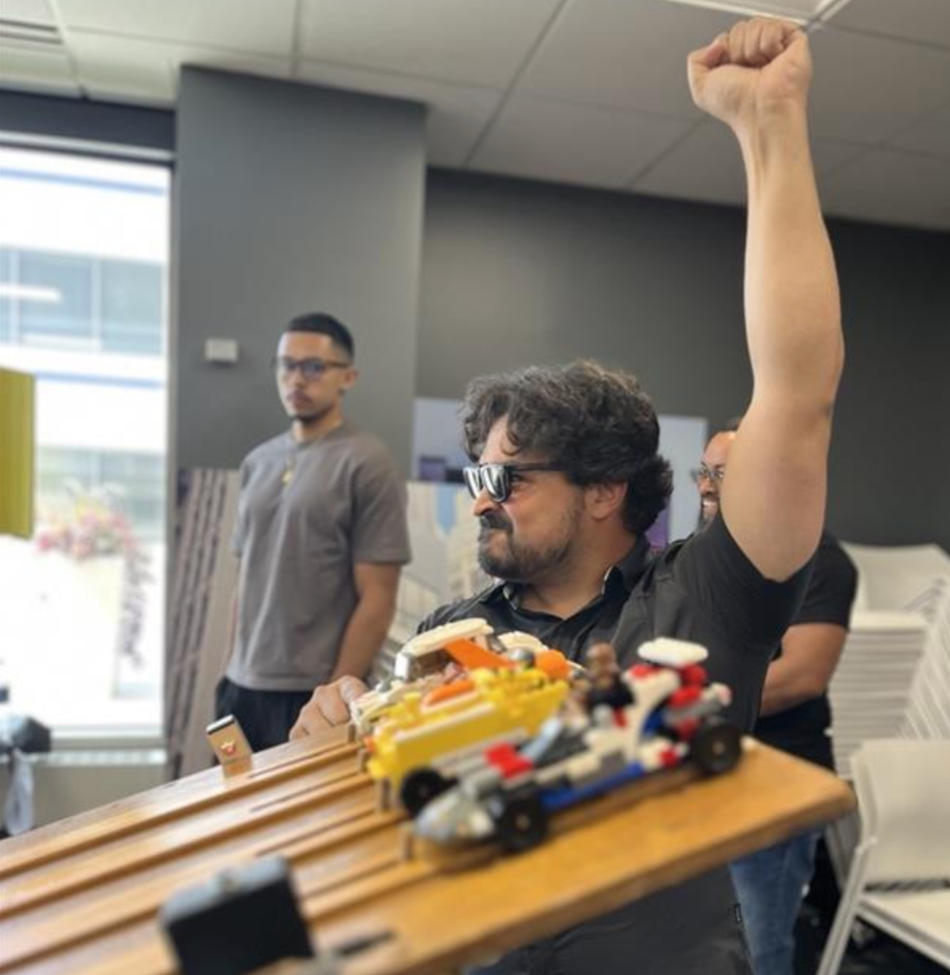 A man wearing sunglasses and a black shirt enthusiastically raises his arm in a fist pump at a store, with toy race cars on a wooden surface in front of him. Two other men are in the background, one with glasses looking on.