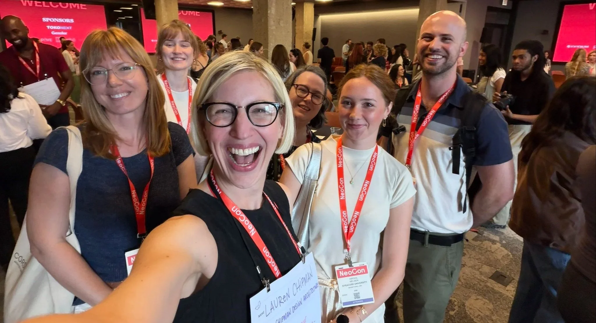 Group of people at a conference or event, smiling for a selfie. They are wearing name tags and lanyards that say "NeoCon." The background shows other attendees and a large red display screen with the word "WELCOME".