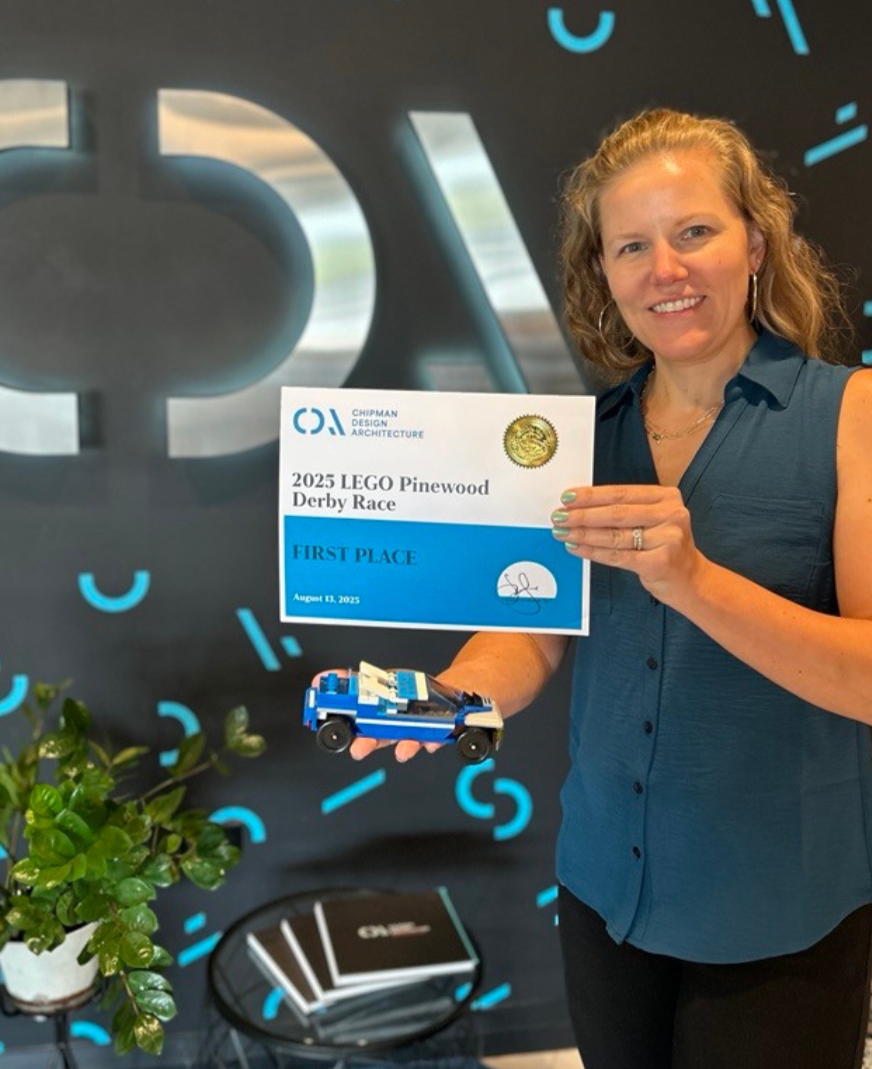 Woman holding a LEGO car and certificate for first place in LEGO Pinewood Derby Race, August 13, 2023, in front of a Chipman Design Architecture backdrop.