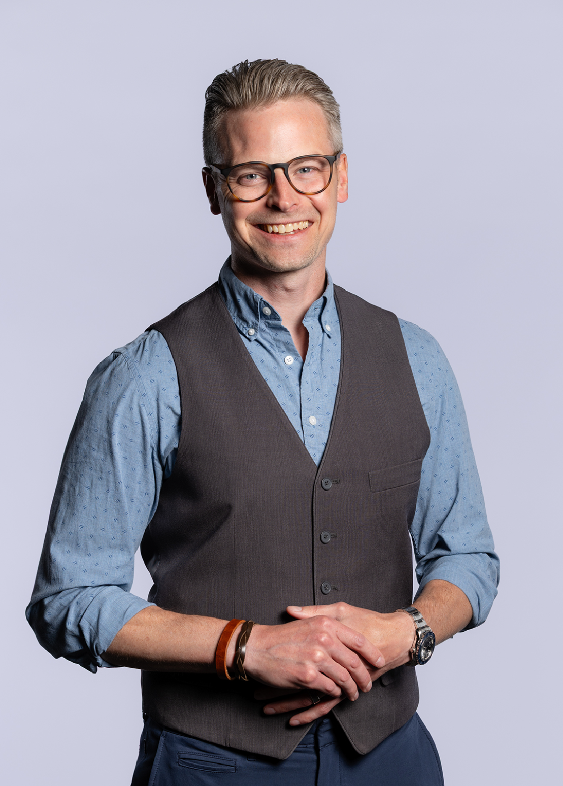 A smiling man with glasses, a blue button-up shirt, a gray vest, and wearing a watch, standing against a plain light background.
