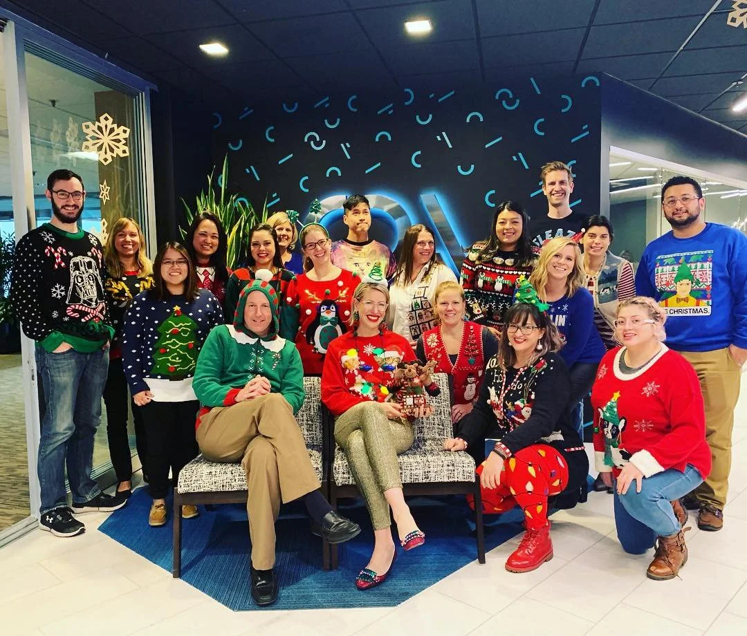 Group of people posing together in holiday sweaters in an office lobby with Christmas decor.