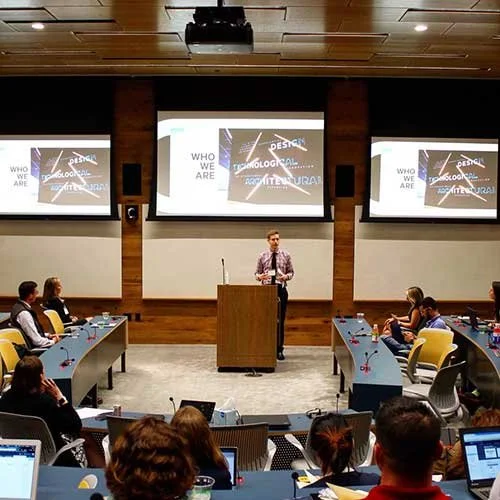 A person presenting at a conference with three large screens behind them displaying a presentation slide, and an audience seated at tables.