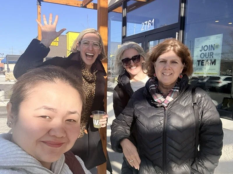 Four women standing outside a storefront, smiling and enjoying a sunny day. One woman in front is taking a selfie, and others are standing behind her, smiling and waving.