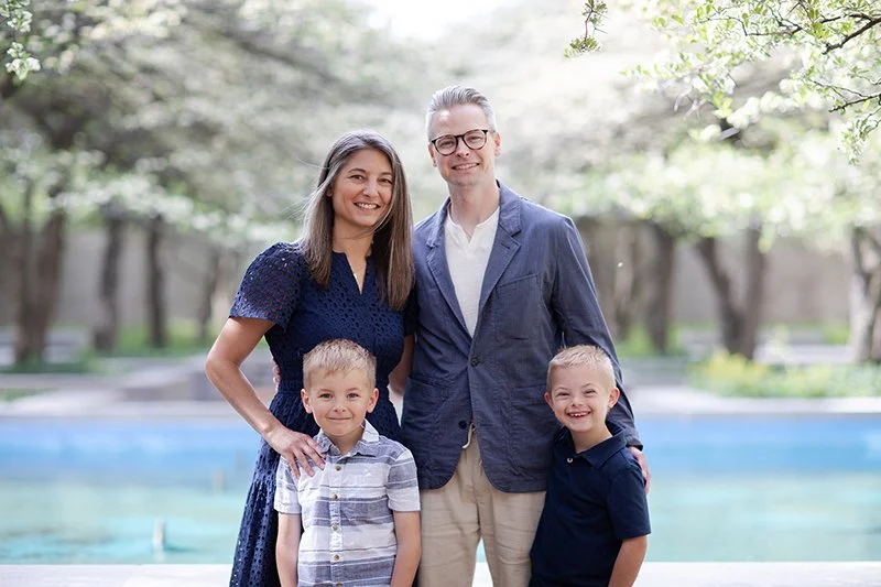 A family of four standing outdoors near a pool, smiling. The family consists of a woman, a man, and two young boys, all dressed casually.