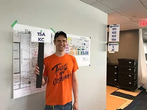 A young man smiling and holding a large black foam prop with a green tape at the top, standing in a hallway near a large floor plan on the wall and directional signs.
